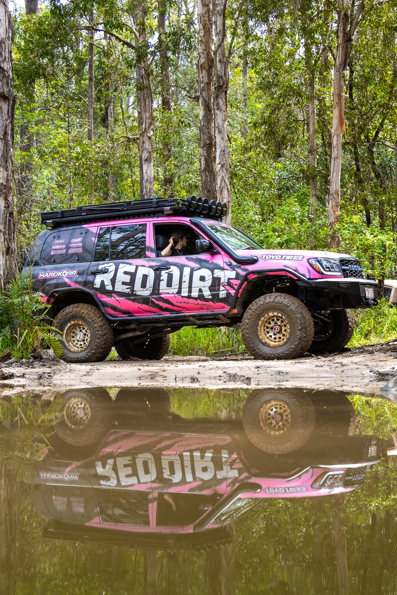 A pink and black off-road vehicle with large tires and a roof rack, parked in a forest near a reflective water puddle.
