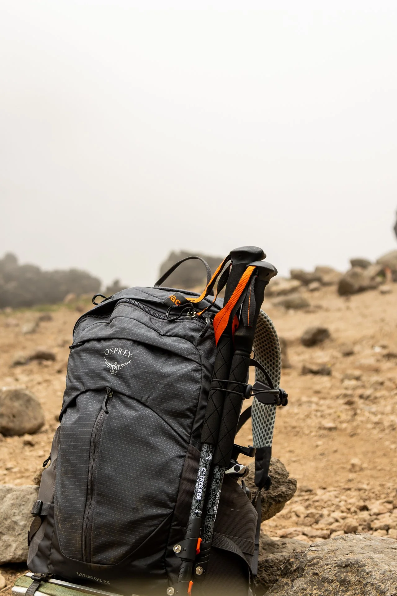 A gray Osprey hiking backpack with a black and orange handle and straps, placed on a rocky terrain with a blurred mountain background.