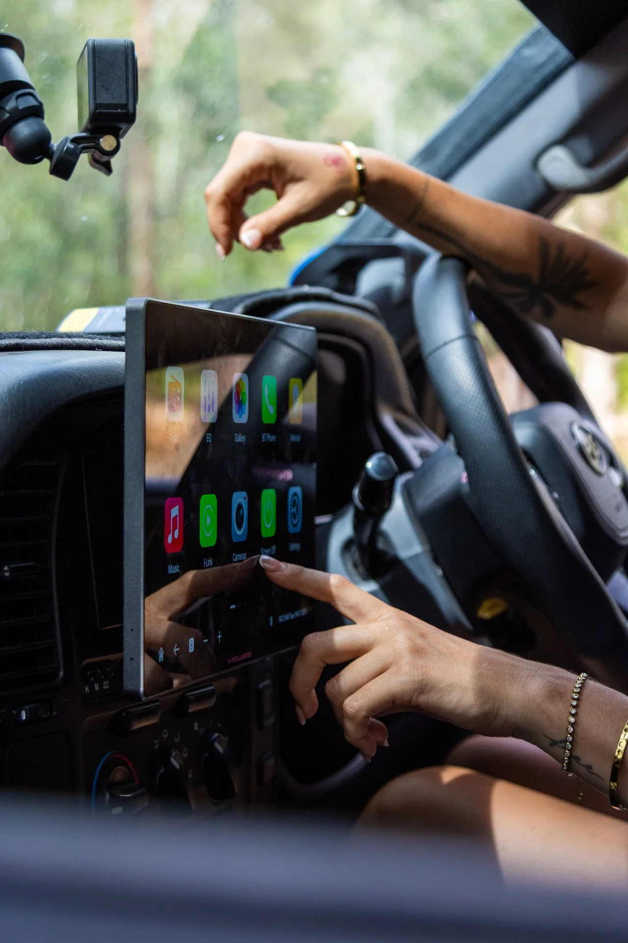 Person in a vehicle using a touchscreen navigation and entertainment system, with a hand adjusting settings and another hand pointing at the screen. The vehicle's dashboard and steering wheel are visible, and the background outside the windshield sho