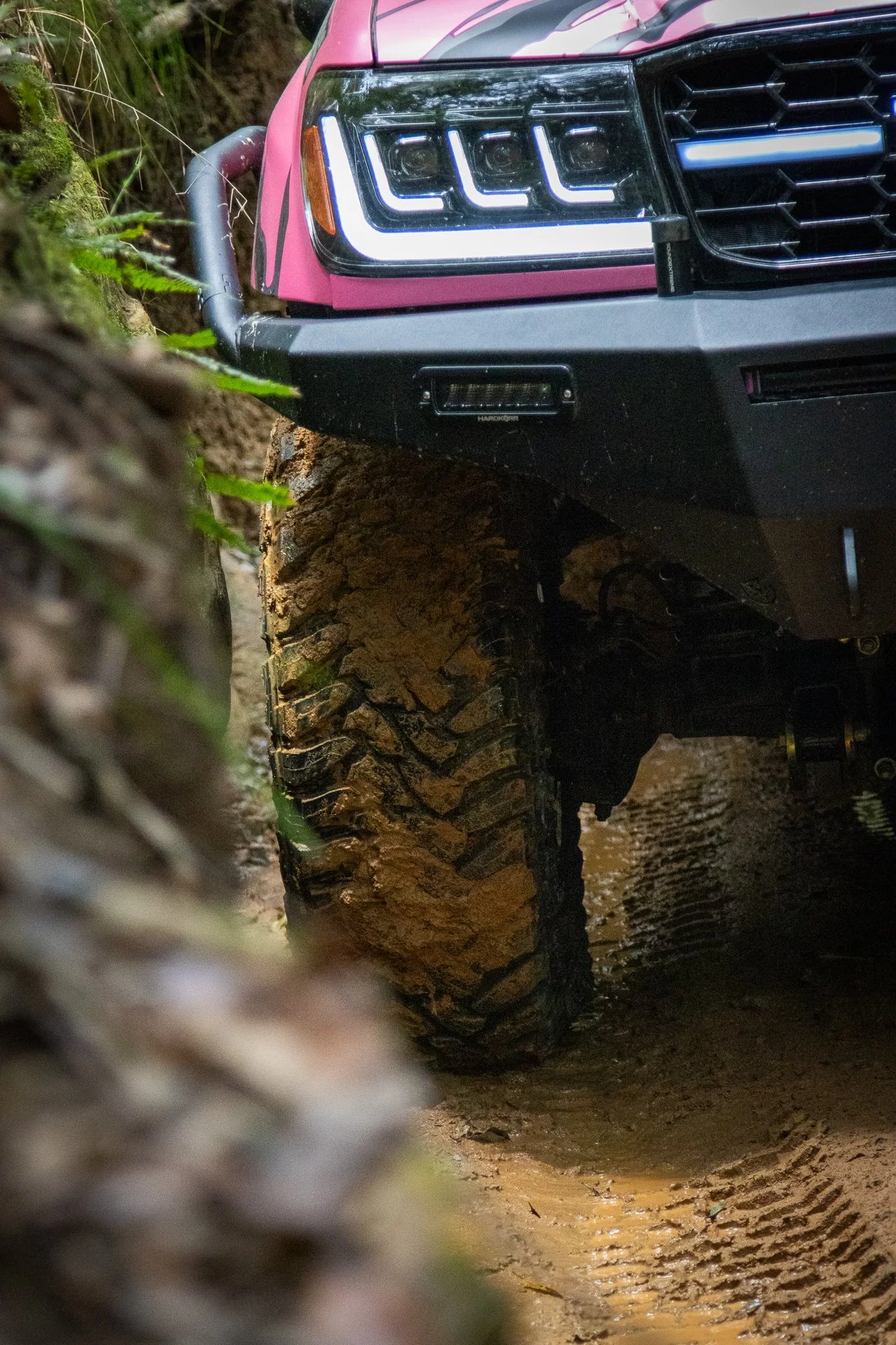 A pink off-road vehicle with large, muddy tires navigating through a muddy trail in a forest.