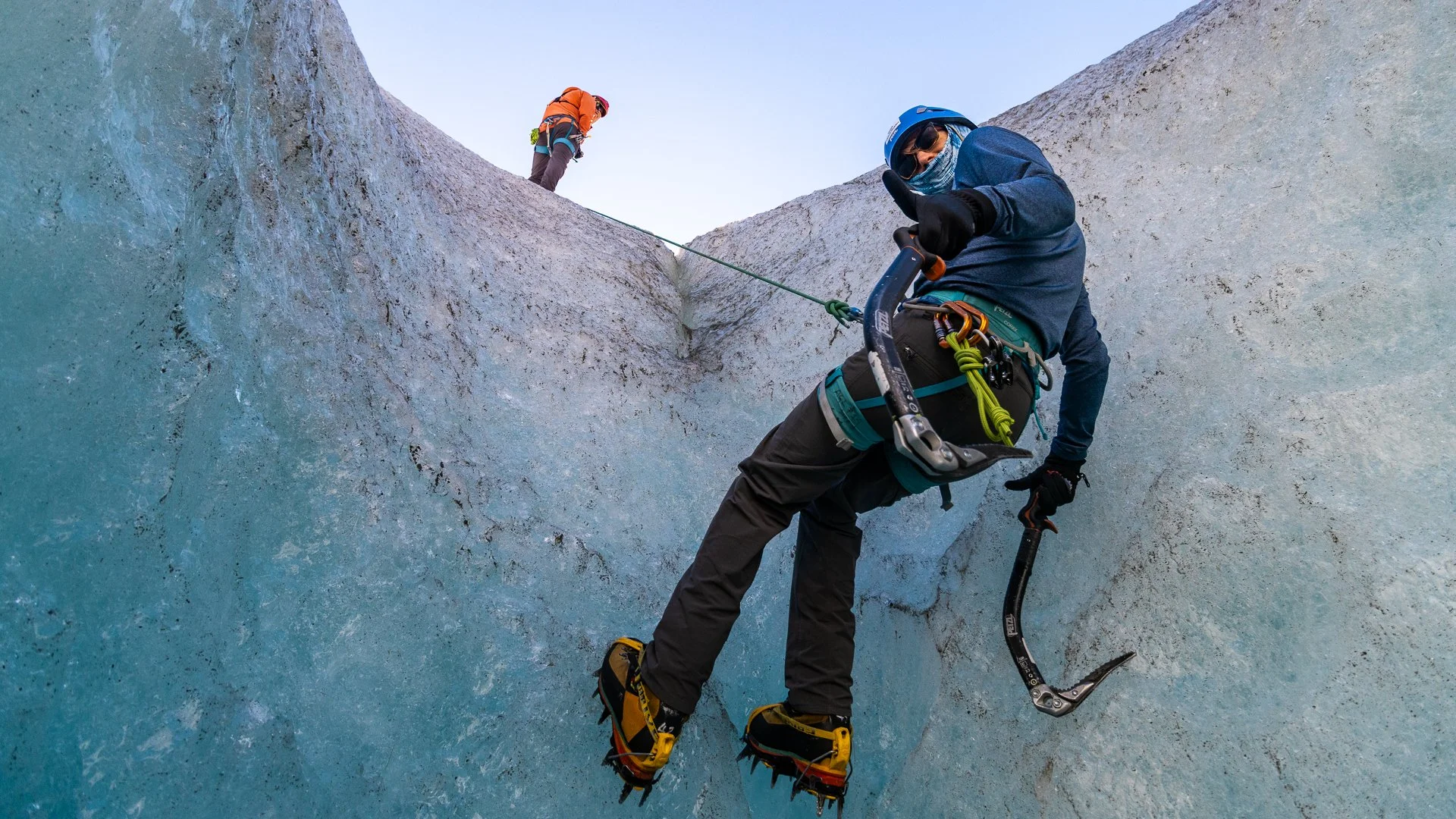 Two climbers ascending a steep ice wall with ice axes and crampons, wearing helmets and outdoor gear.