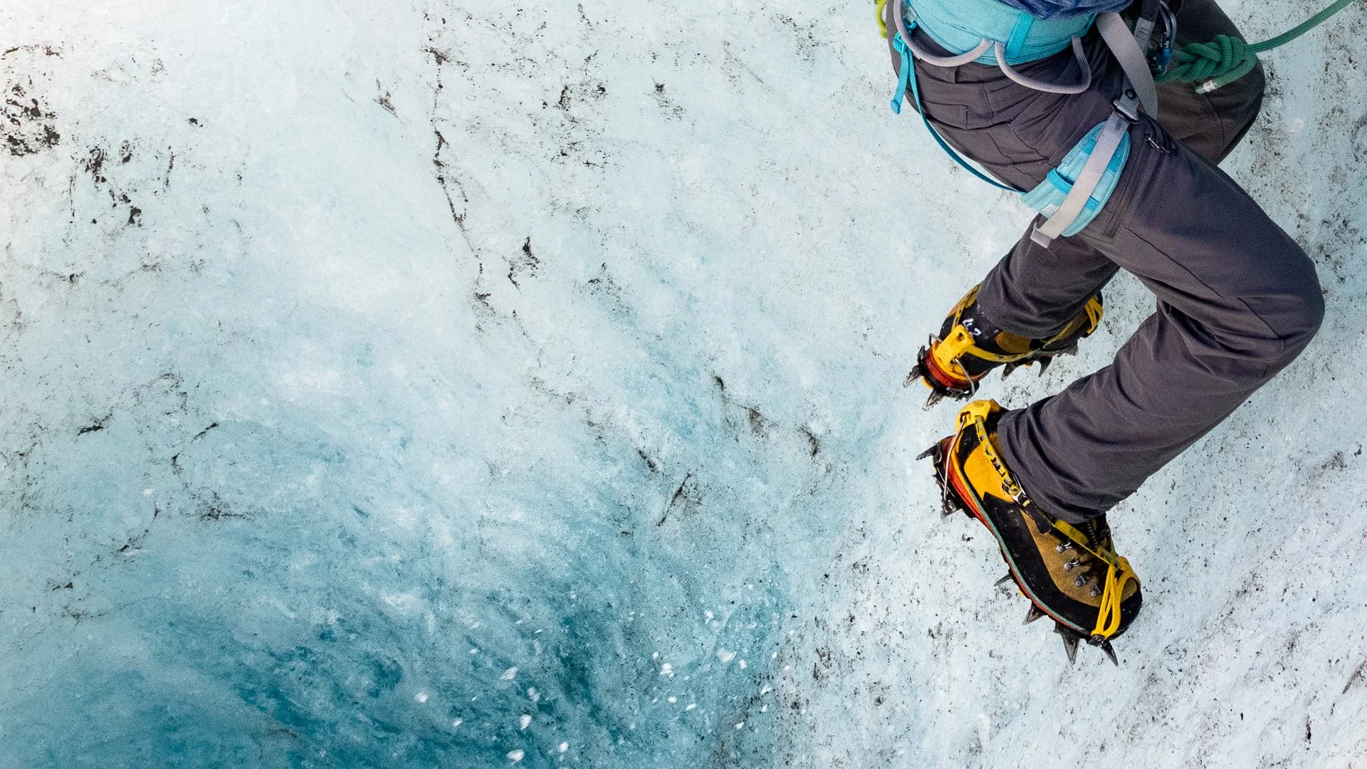 Close-up of a person wearing climbing boots, scaling an icy surface with ice axes, gear, and safety equipment.
