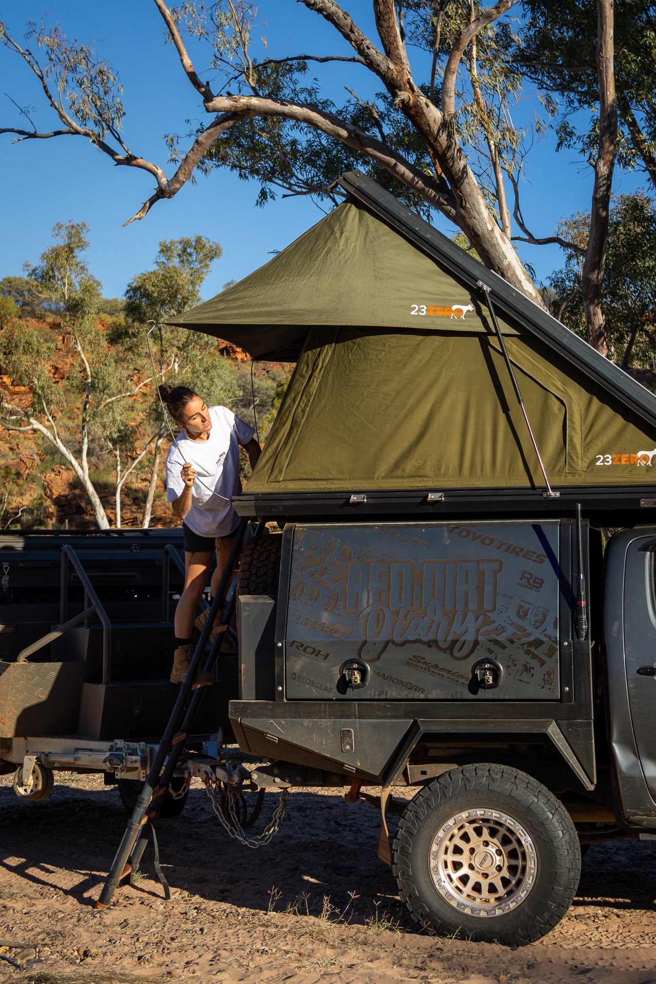 A woman setting up a rooftop tent on a black off-road truck in a desert landscape with trees and blue sky.