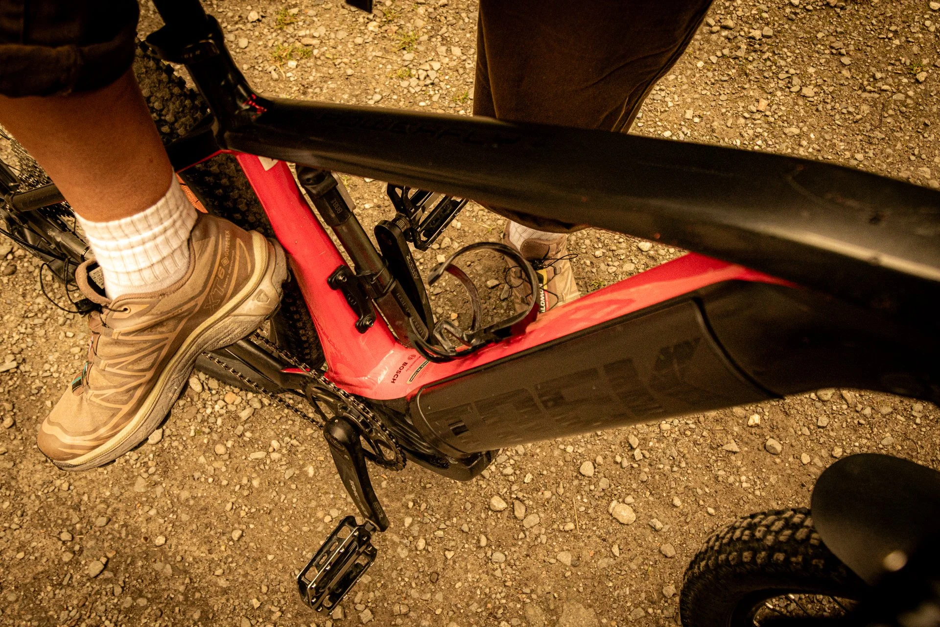 Overhead view of a person wearing beige hiking shoes, black pants, and white socks, standing next to a red electric bicycle on a gravel surface.