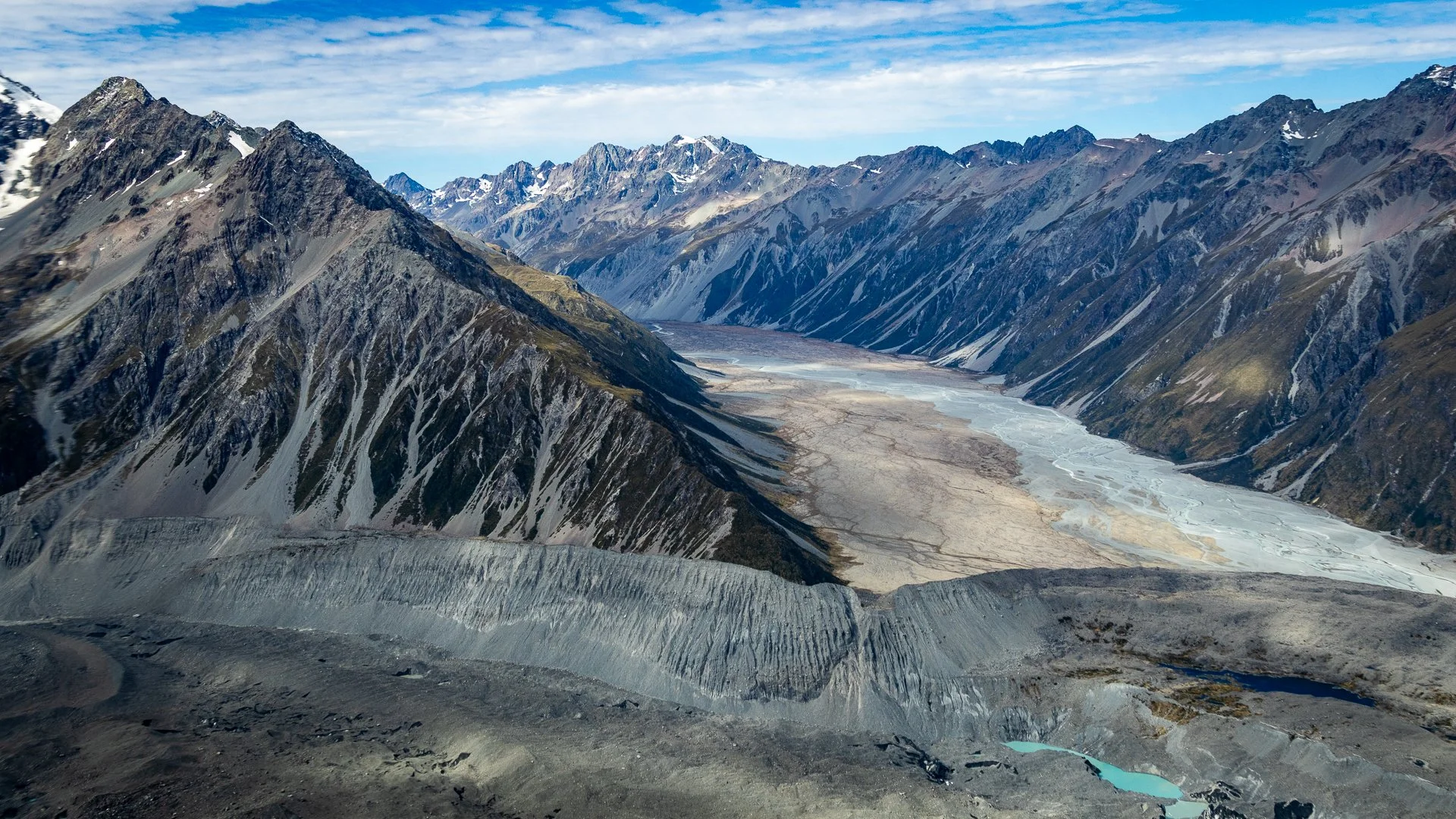 Snow-capped mountains with rocky slopes and a dry riverbed in a high-altitude landscape under a partly cloudy sky.