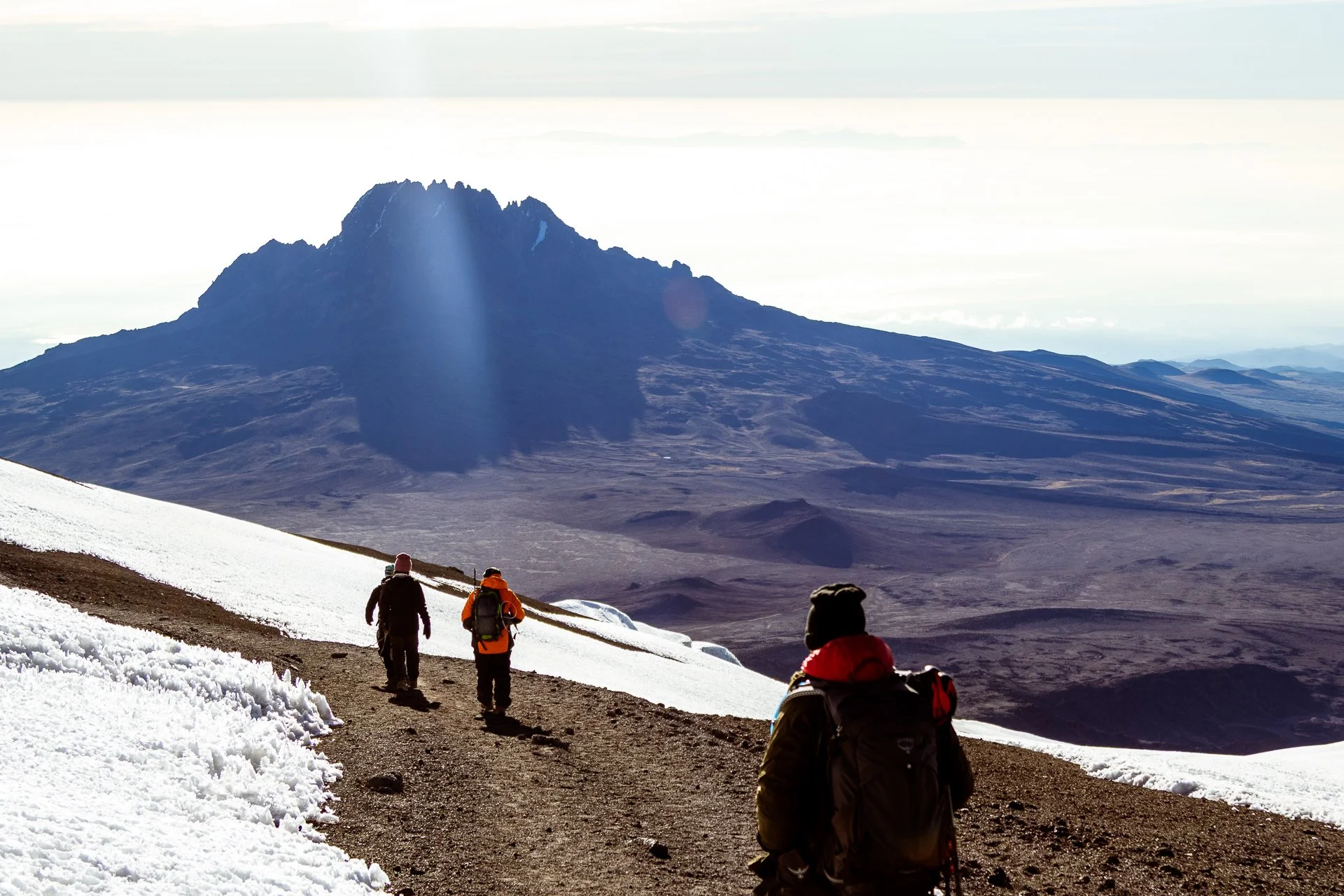 Four hikers walking on a snow-covered trail, with a volcanic mountain in the background under a cloudy sky.