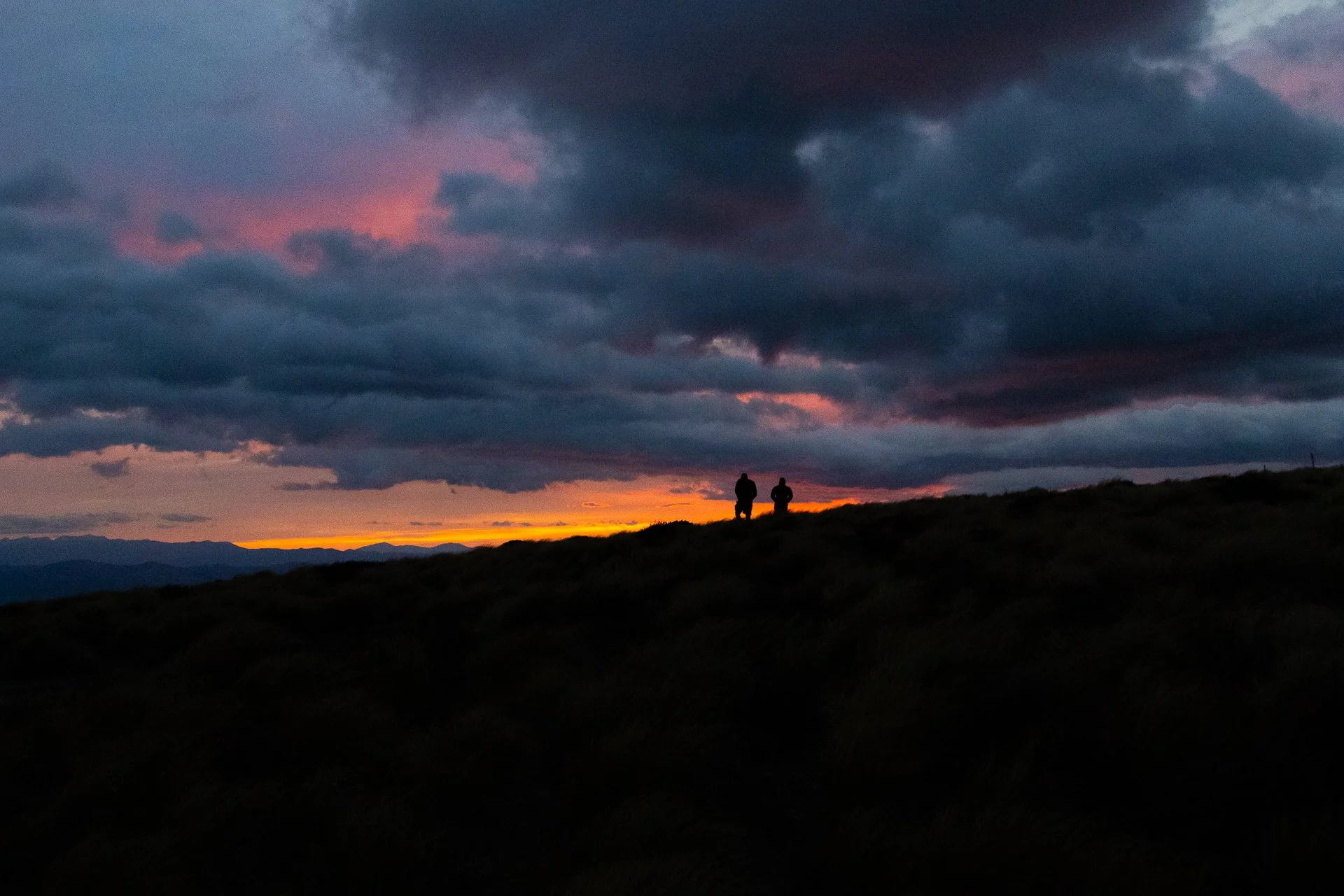 Two people walking on a hill at sunset under a cloudy sky with dark clouds and hints of pink and orange light.