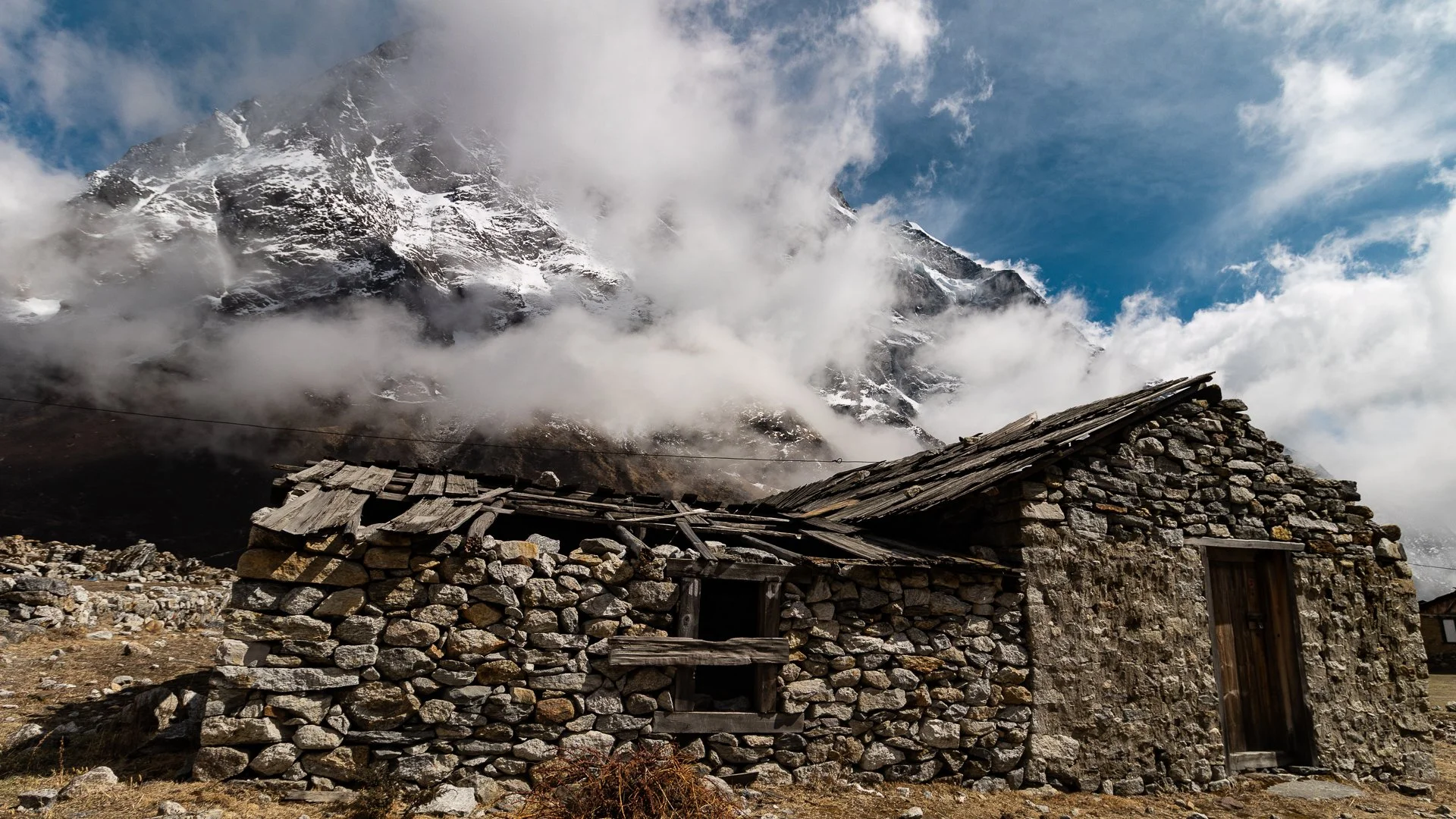 A stone cabin with a wooden roof in front of snow-capped mountains partly covered by clouds.