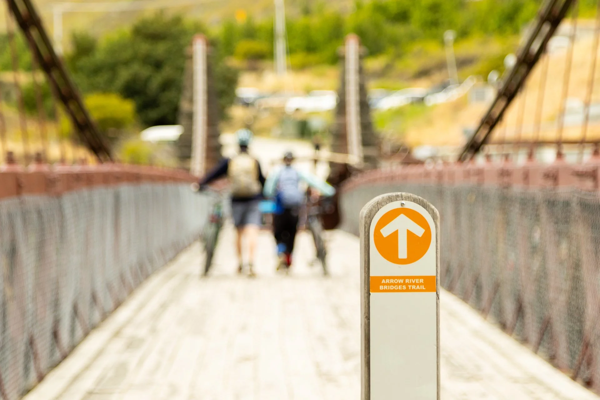 A bridge on the Arrow River Bridges Trail with a sign in the foreground indicating the trail. Two people are walking their bikes across the bridge, with trees and cars visible in the background.