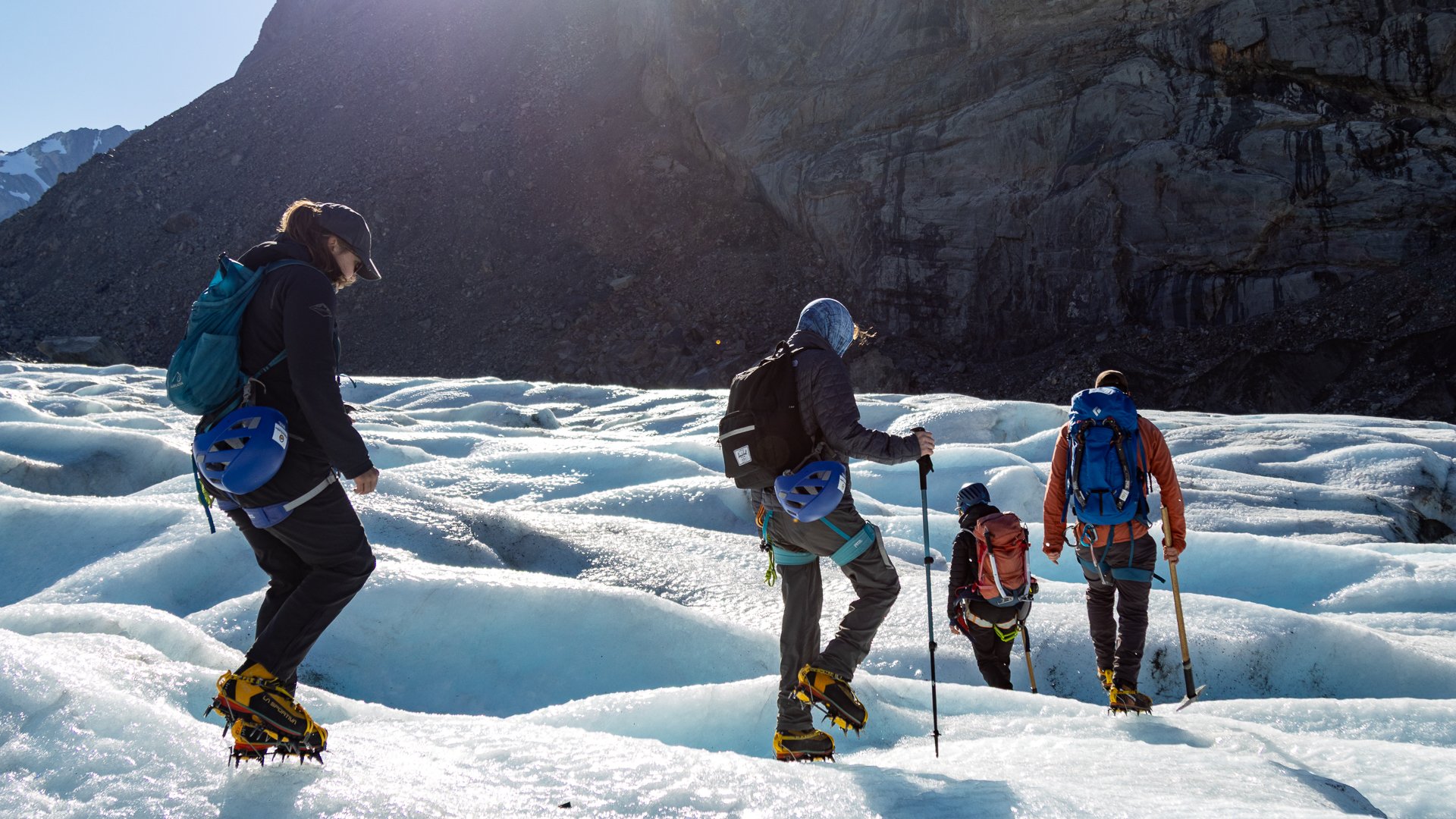 Four people hiking on snow-covered terrain with ice cleats, all carrying backpacks and using trekking poles, surrounded by rugged mountain landscape.