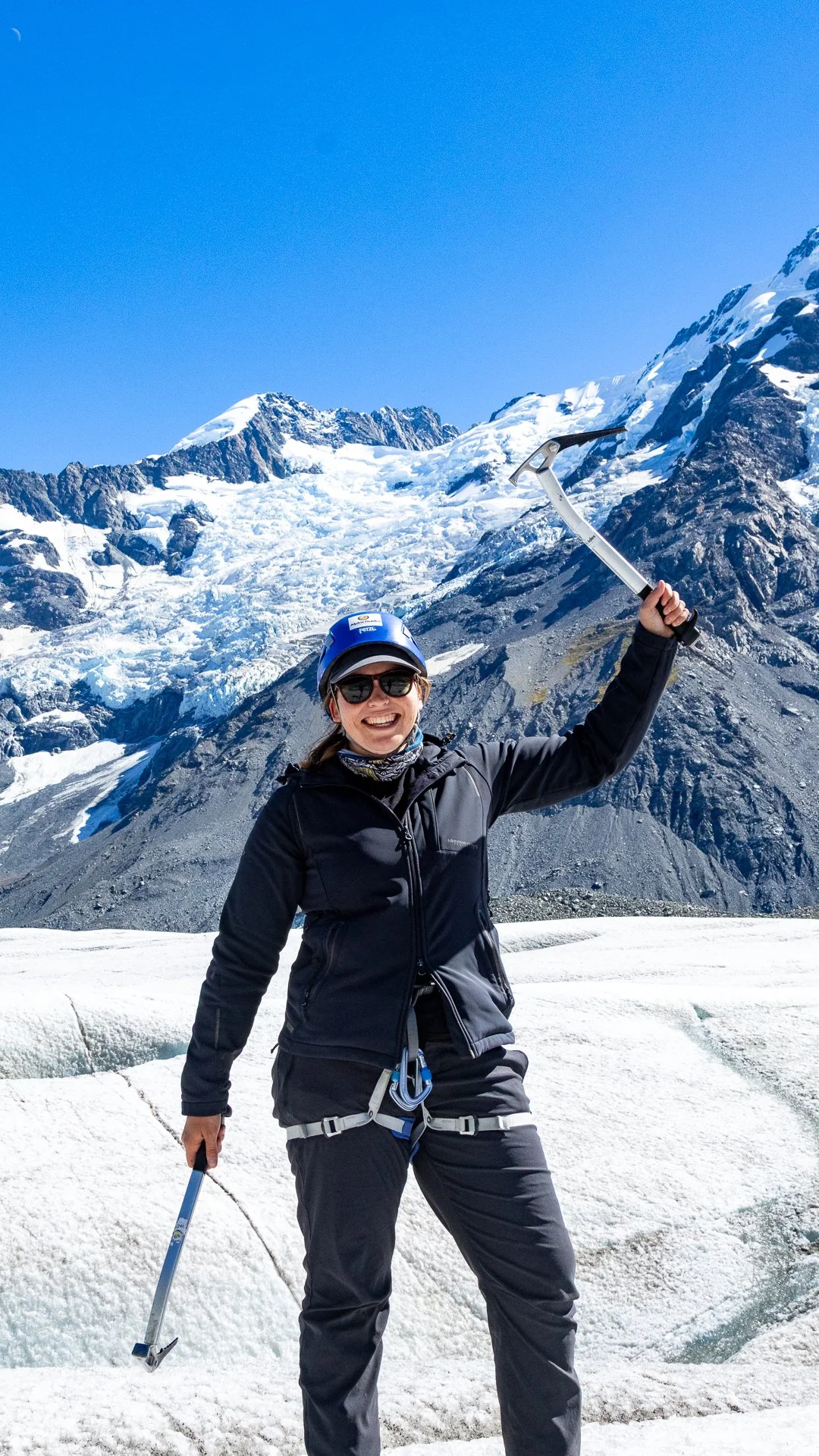 A woman dressed in black outdoor gear, wearing sunglasses and a helmet, holding an ice axe, standing on snow with a mountainous glacier behind her under a clear blue sky.