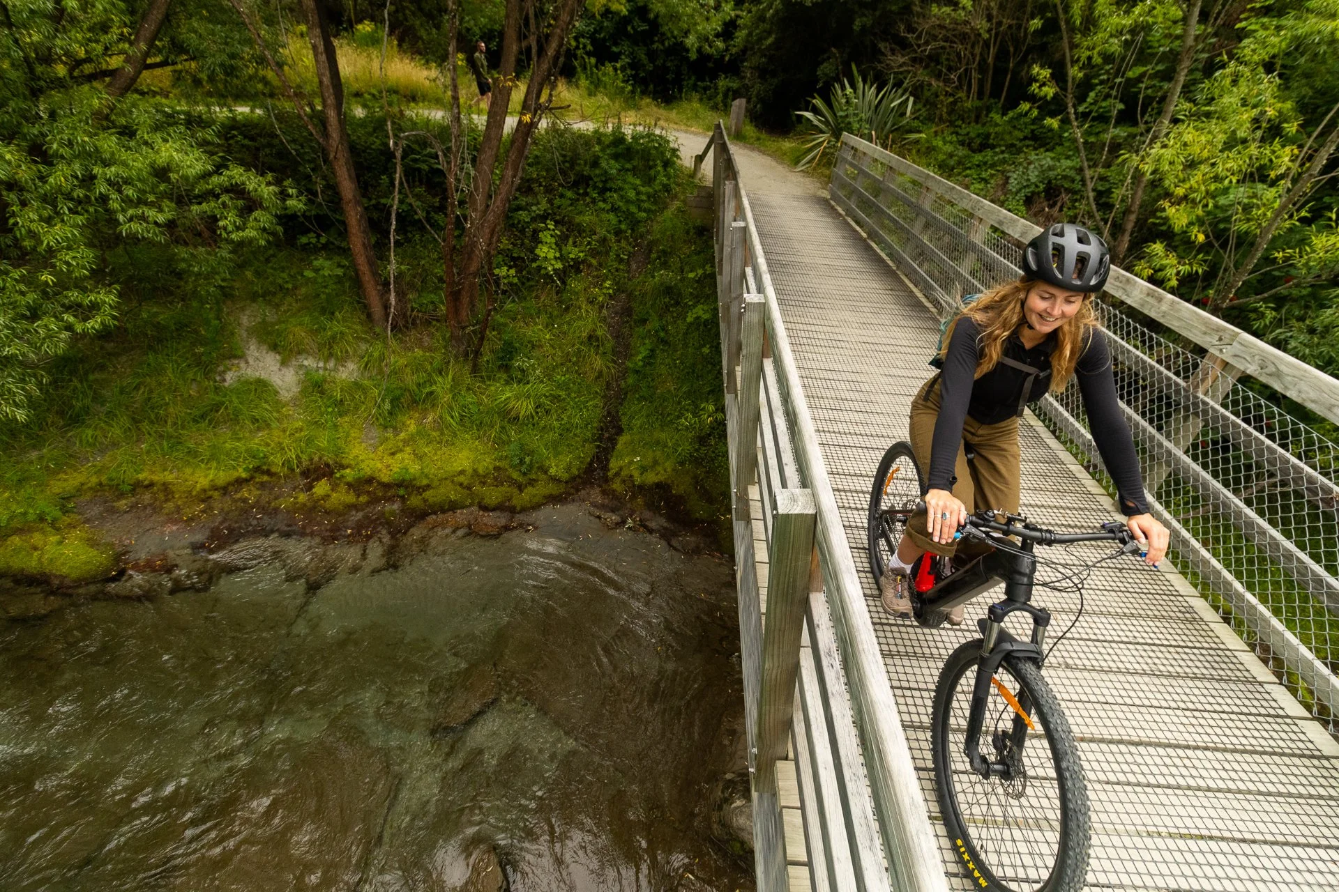 A woman wearing a black helmet and black long sleeve shirt rides a bicycle across a metal bridge over a creek, surrounded by green trees and foliage.