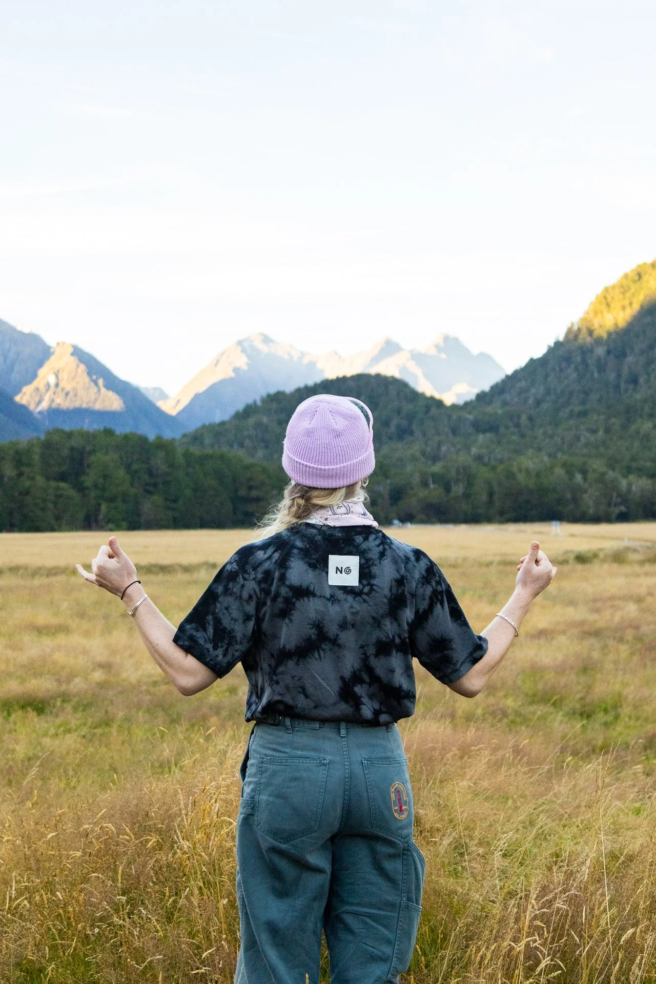 Person standing in a field with mountains in the background, wearing a pink beanie, a black tie-dye shirt, and blue jeans, facing away, with arms raised in a meditative pose.