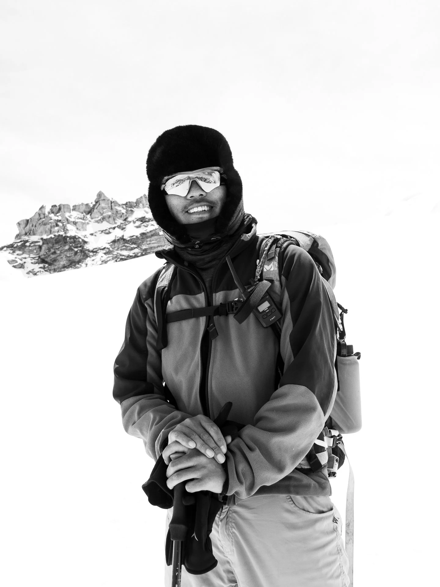 Black-and-white photo of a person wearing a winter hat, ski goggles, a jacket, and backpack, standing outdoors in a snowy mountain landscape.
