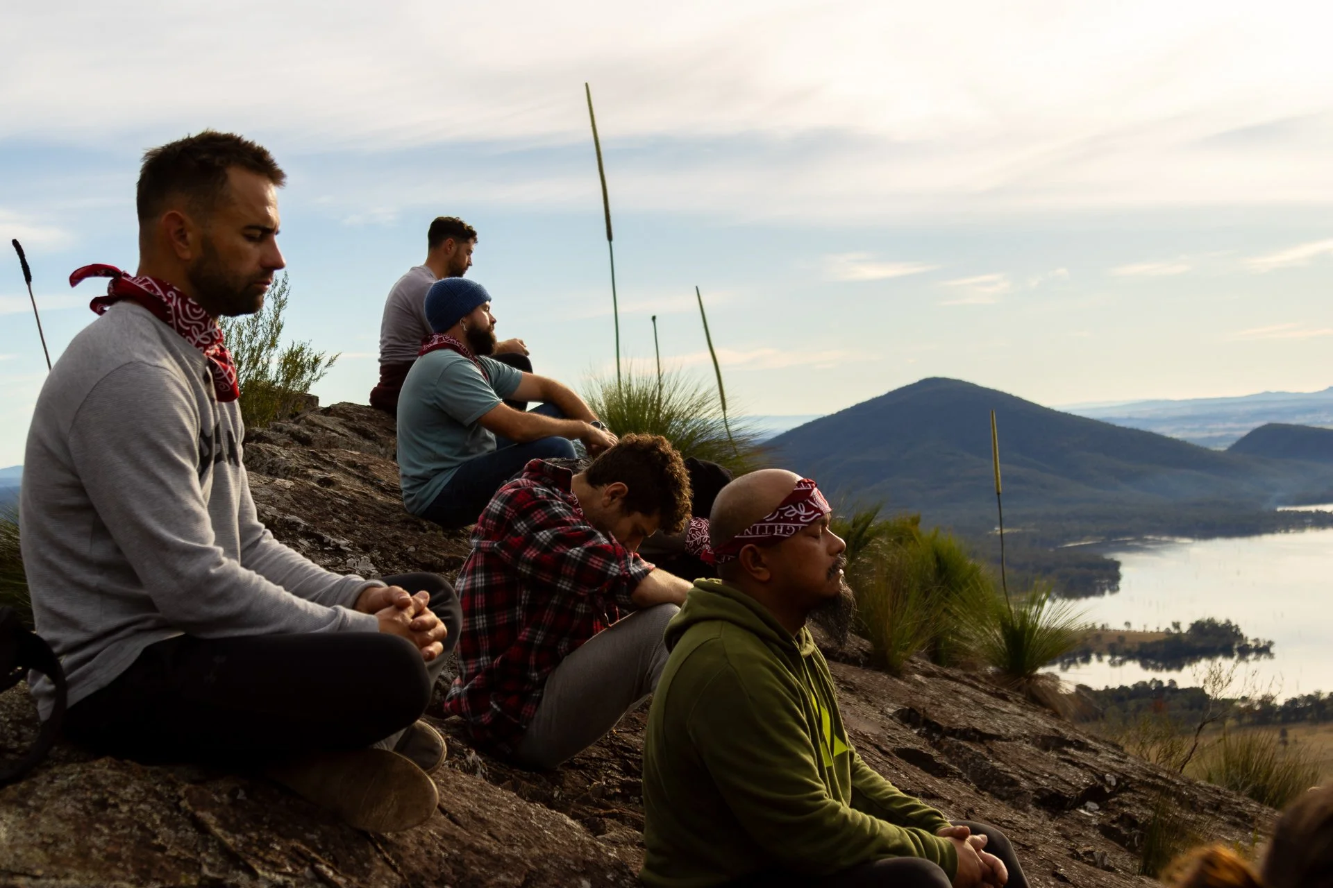 Group of men meditating outdoors on a hillside overlooking a river and mountains, some wearing bandanas and casual clothing, sitting cross-legged with eyes closed and hands resting on knees.