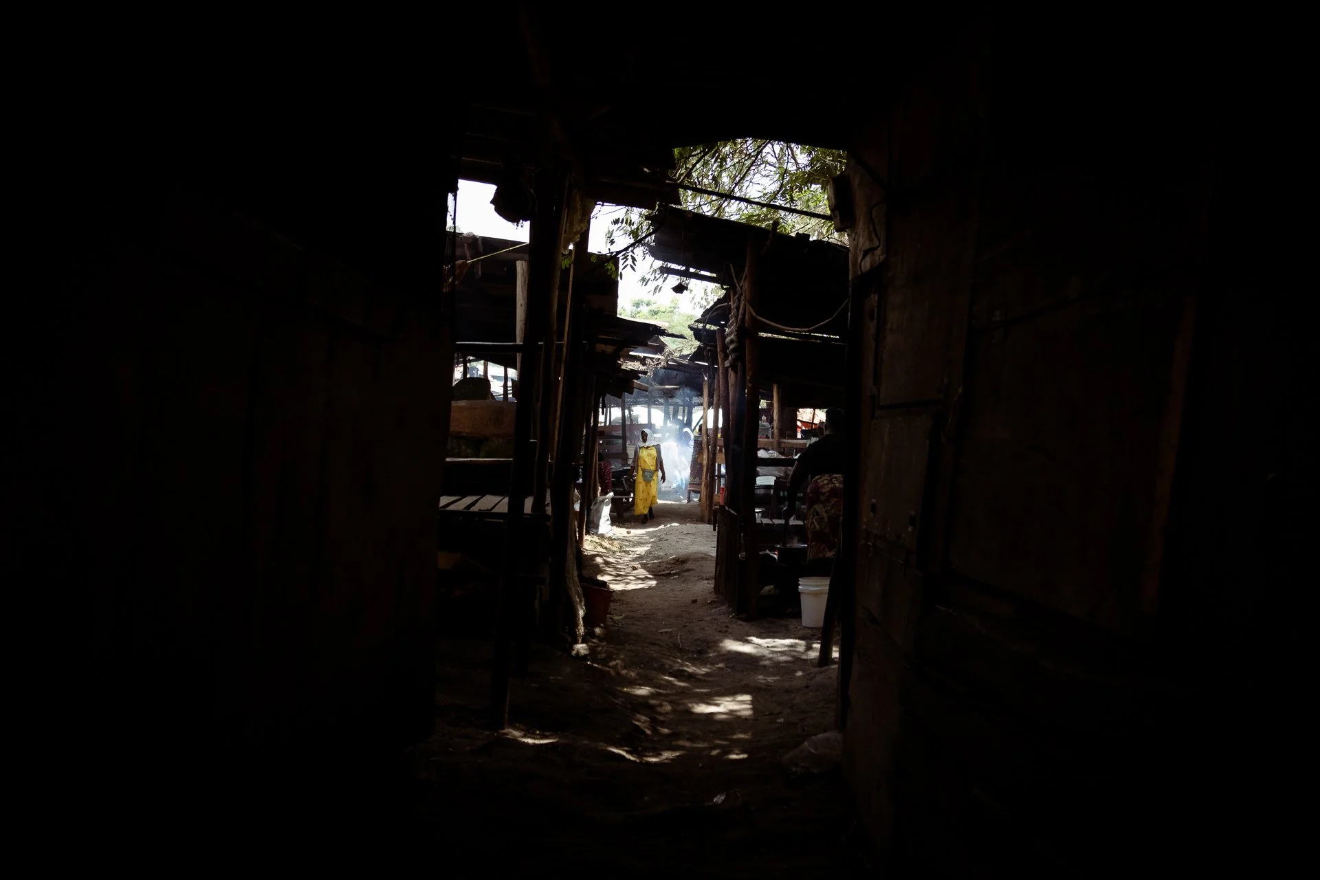 View through a dark, wooden doorway showing people walking outside in a rural area with makeshift structures and trees.