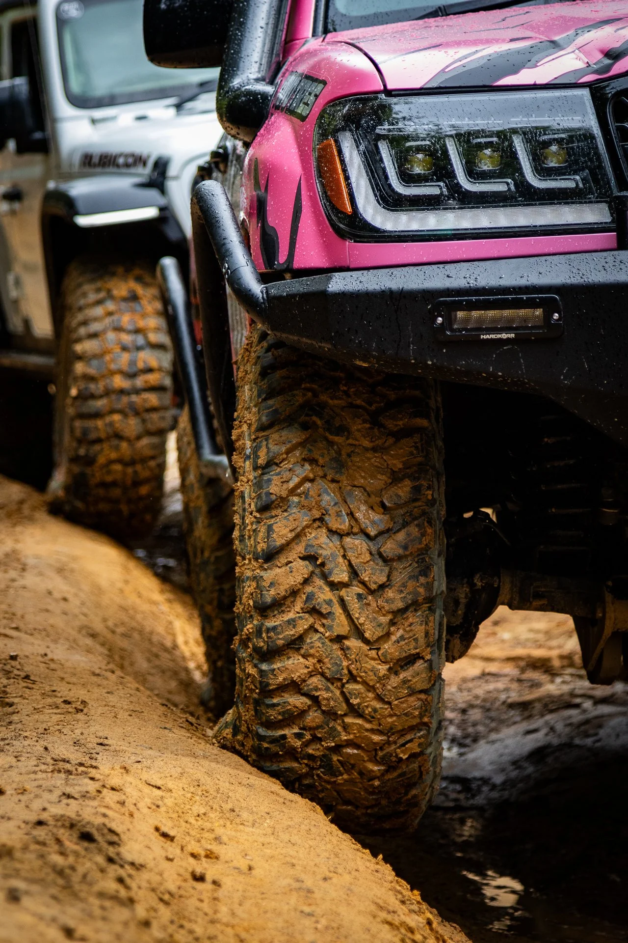 Close-up of mud-covered off-road tires on a pink and black Jeep vehicle, with water droplets on the headlight, on a dirt trail.