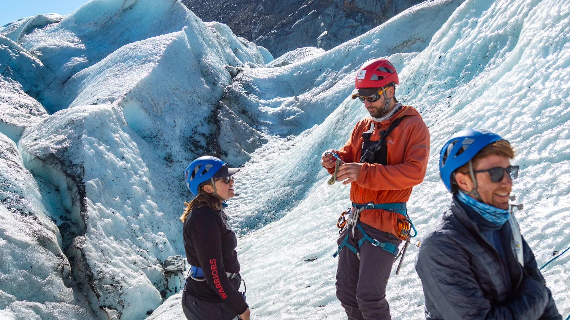 Three people in helmets and outdoor gear standing on snow-covered ice with glaciers in the background, one man is adjusting equipment while two women smile.