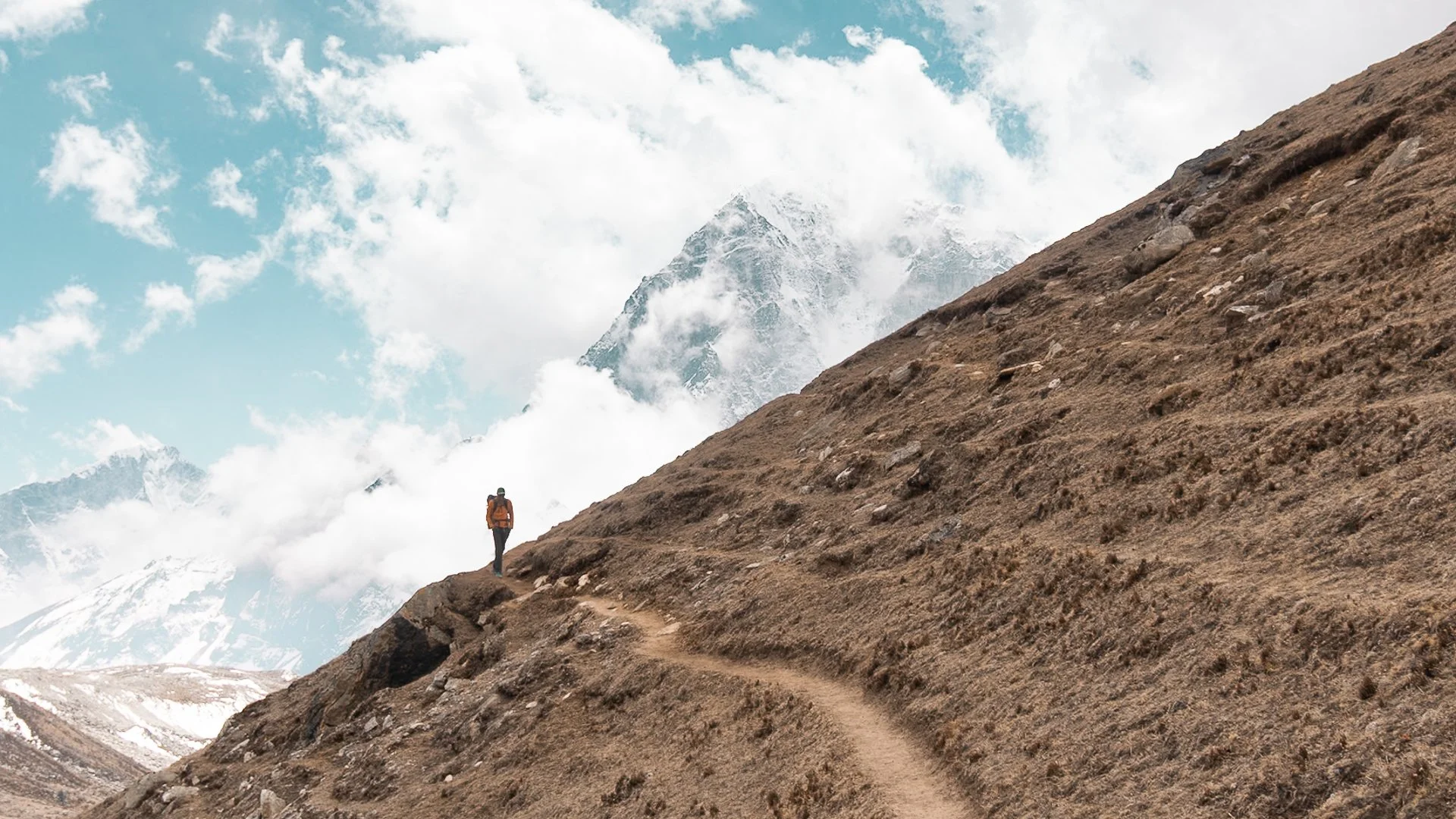 A hiker walking along a narrow mountain trail with snow-capped peaks and clouds in the background.
