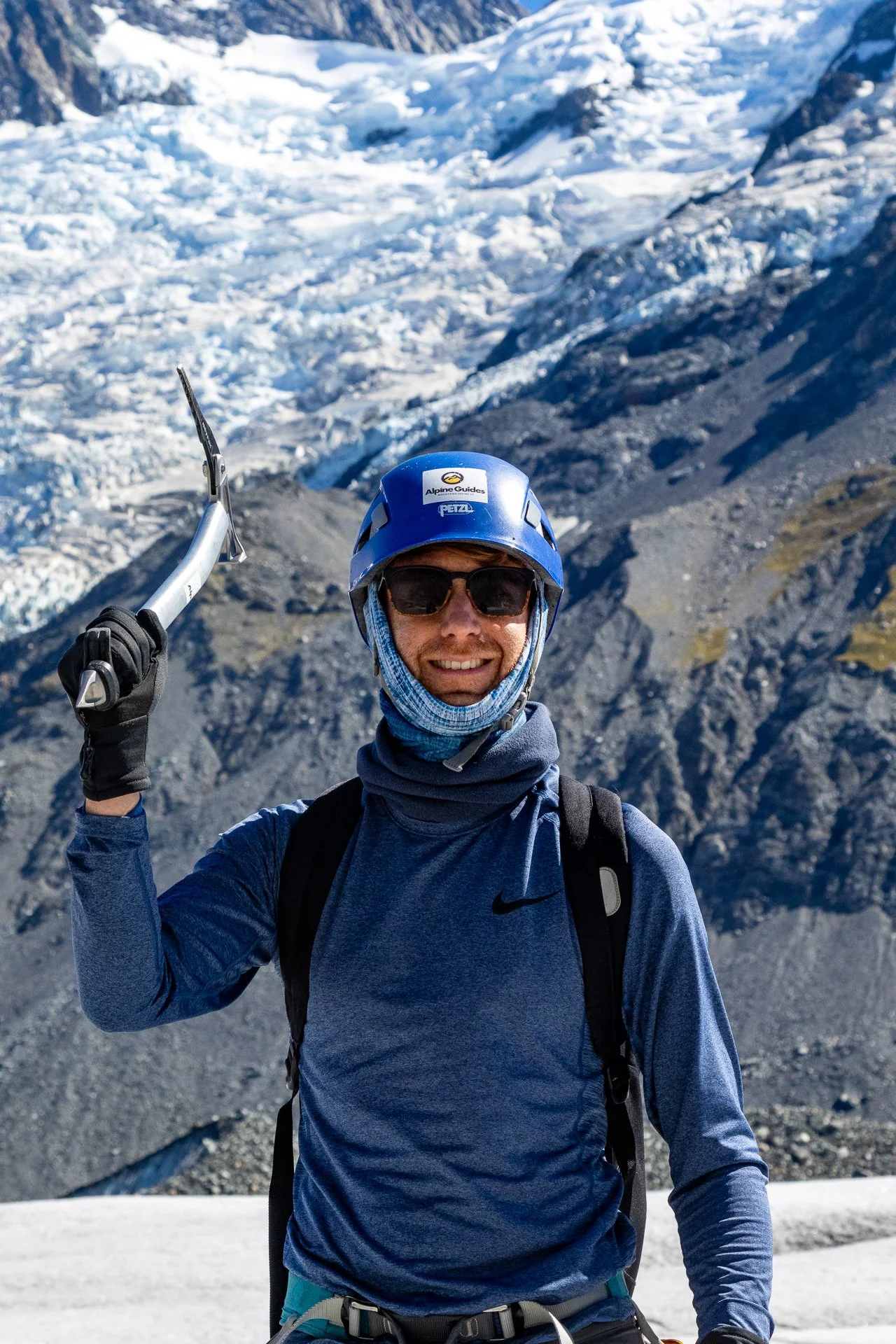 A man smiling in outdoor snowy mountain terrain, wearing a blue helmet with a logo, sunglasses, a blue long-sleeve shirt, and carrying a backpack.
