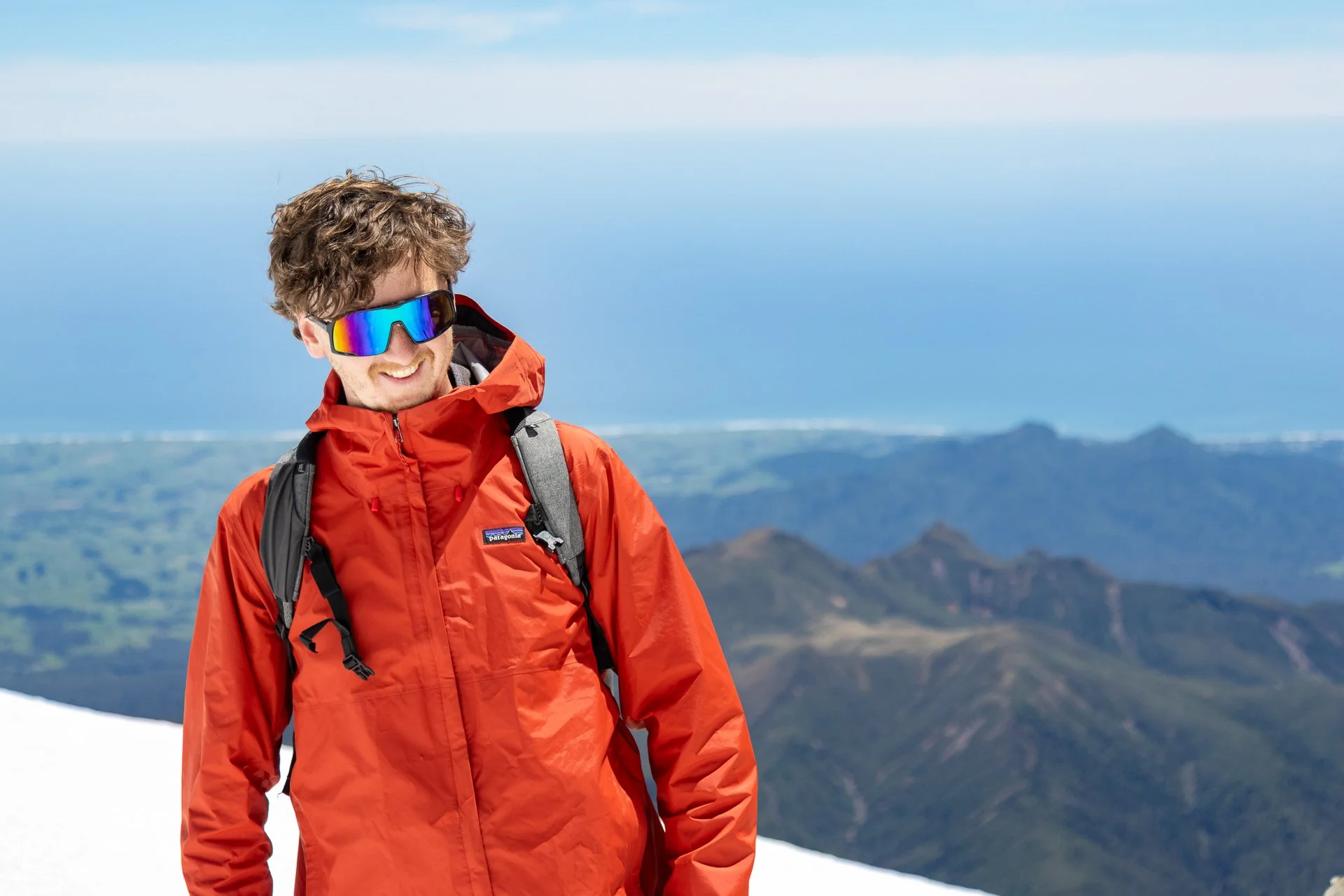 A man wearing a red outdoor jacket, colorful reflective sunglasses, and a backpack, standing on a snowy mountain with scenic green hills and distant mountains in the background.