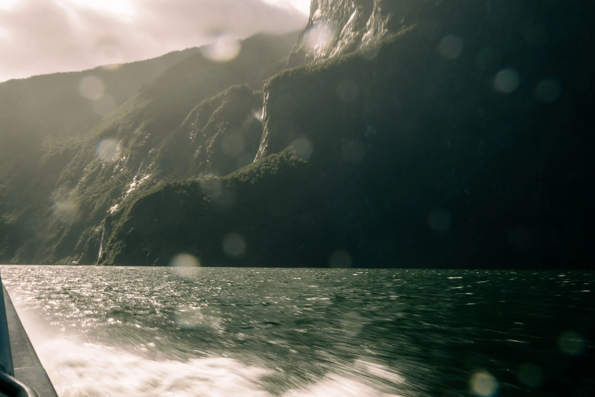 A view of a mountainous shoreline with steep, green hills and cascading waterfalls, taken from a boat on the water.