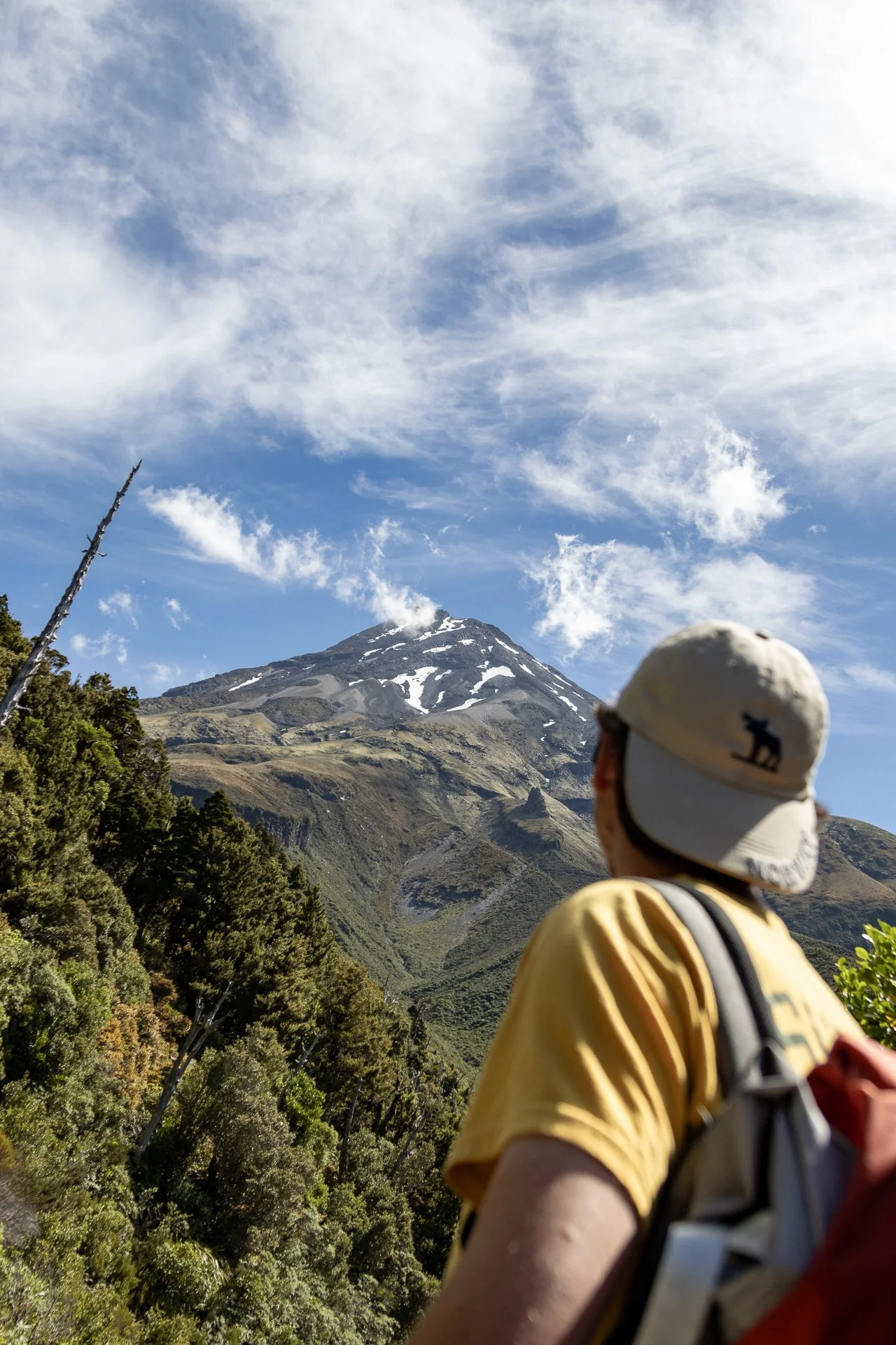 A person wearing a beige cap with a bear logo, a yellow shirt, and a backpack is looking at a snow-capped mountain under a blue sky with white clouds.