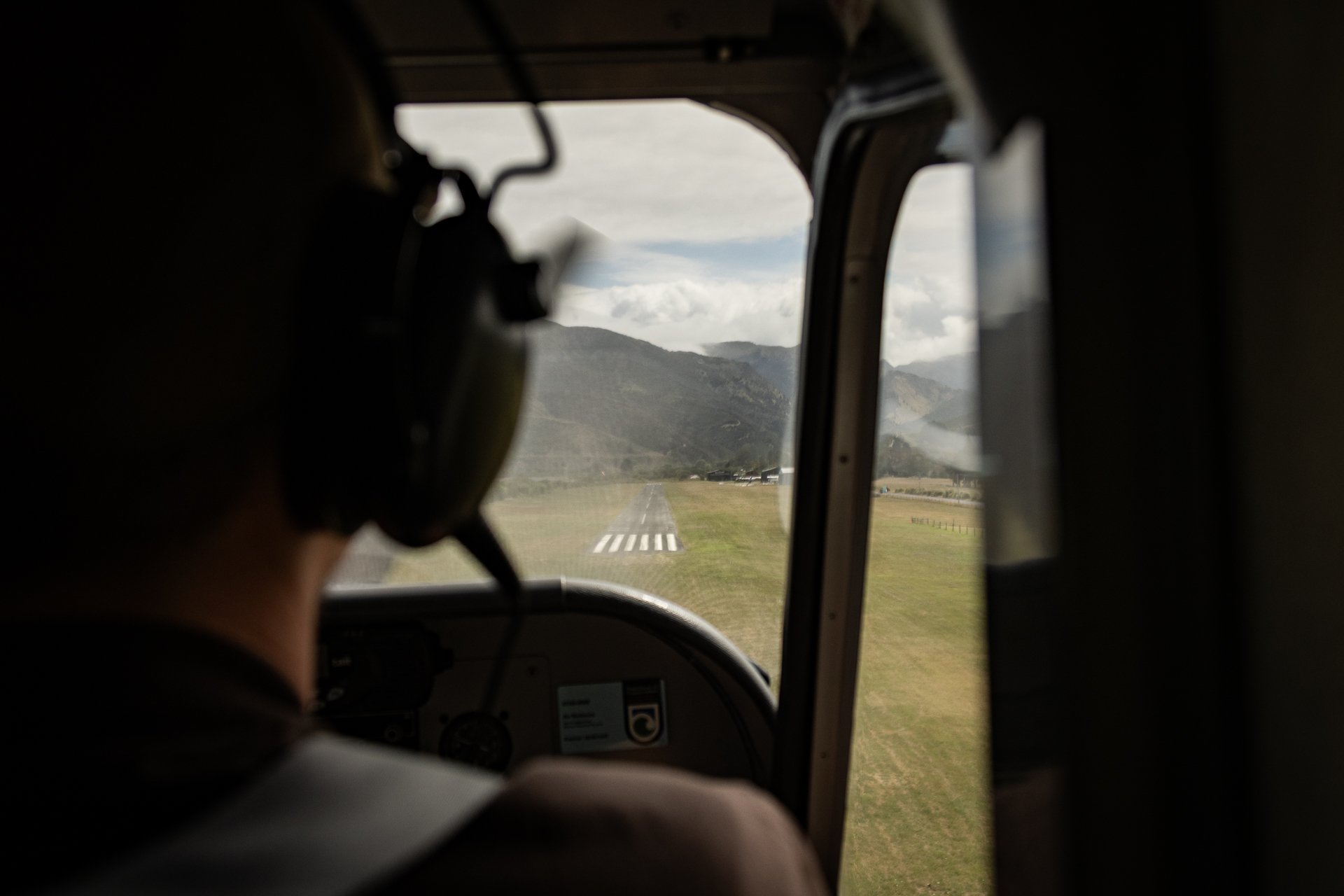 Photo taken from inside a helicopter cockpit showing the pilot's silhouette with mountains and a runway visible through the windshield.