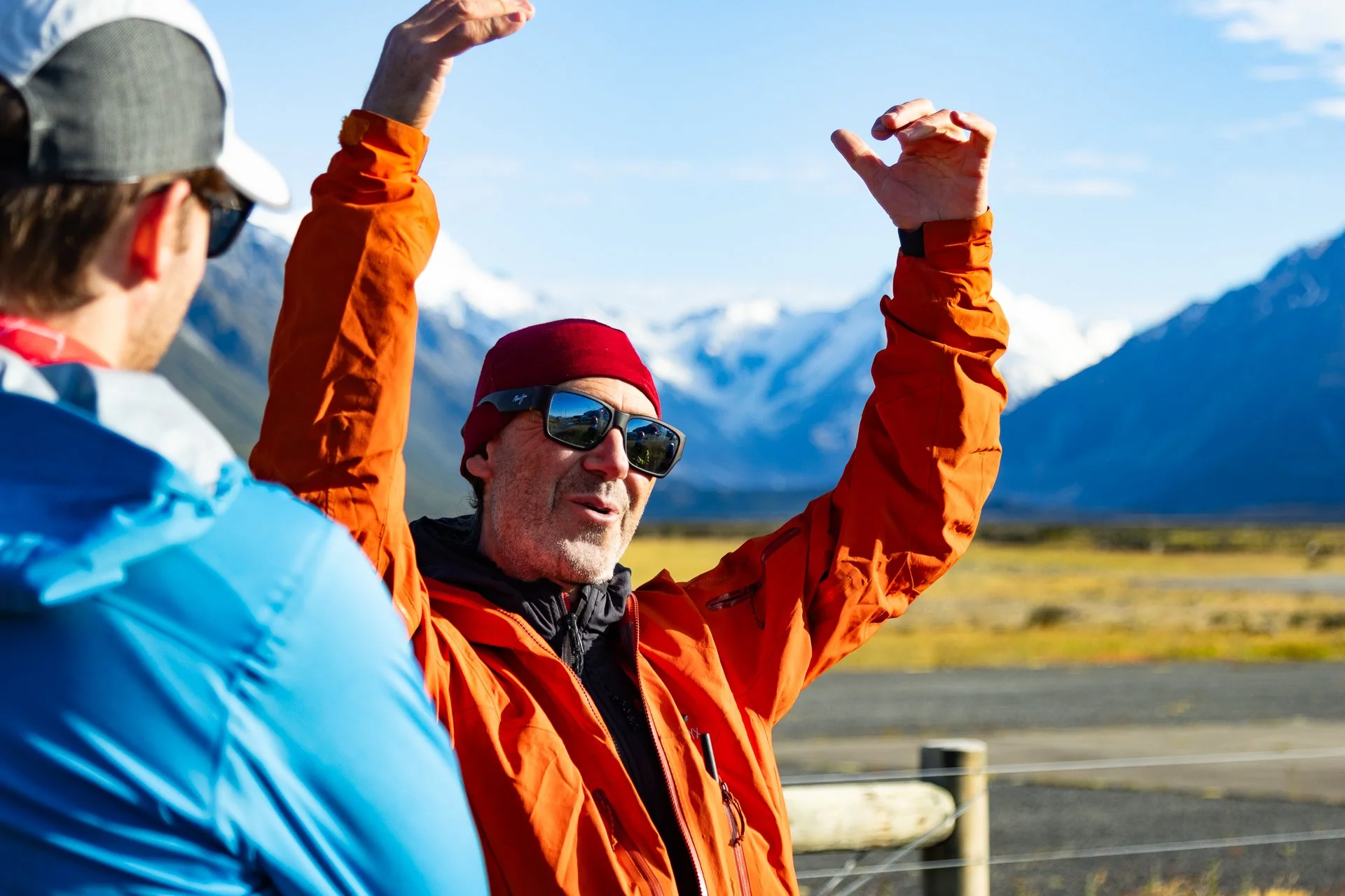 A man in an orange jacket and a red beanie, wearing sunglasses, is raising his hands and speaking to another person in a blue jacket, with snow-covered mountains in the background.