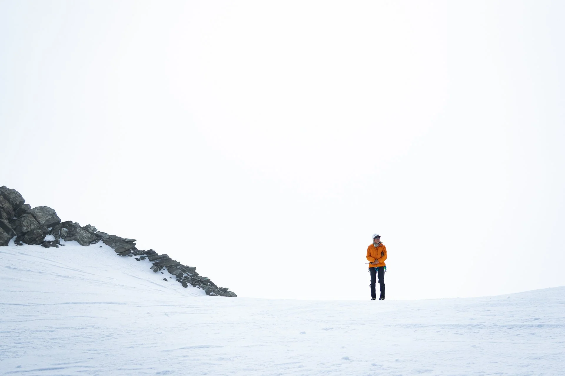 A person standing in a snowy mountain landscape, wearing an orange jacket and a white helmet, with rocks and snow around him under a mostly cloudy sky.