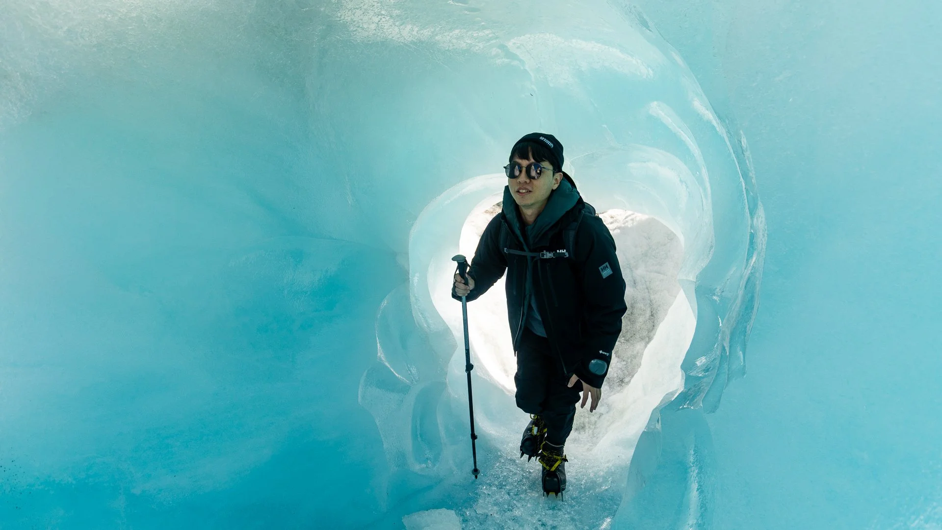 A man in winter gear, wearing sunglasses and a beanie, standing inside an ice cave with blue ice walls, holding an ice axe.