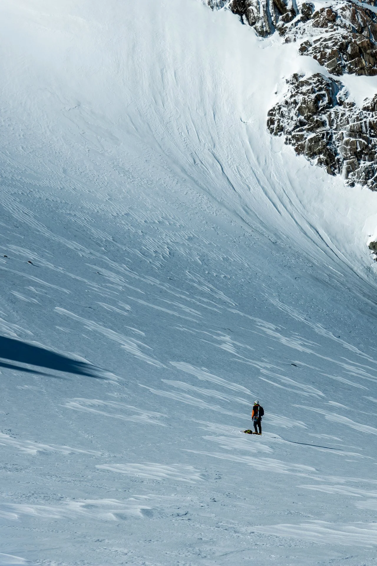 A lone climber standing on a snowy mountain slope beneath a large, snow-covered cliff with rocky outcroppings in the background.
