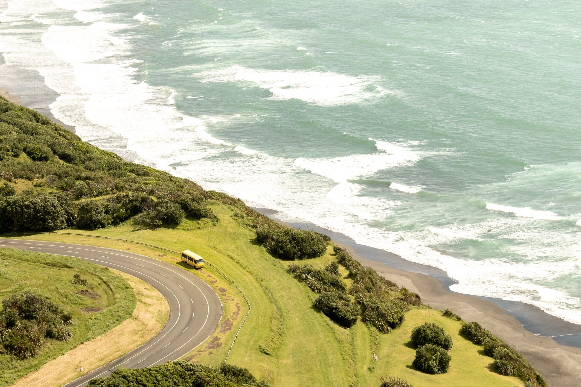 A coastal landscape with a winding road and a bus on a grassy hill, overlooking the ocean with waves crashing on the shoreline, and a green, hilly area covered with bushes and trees.