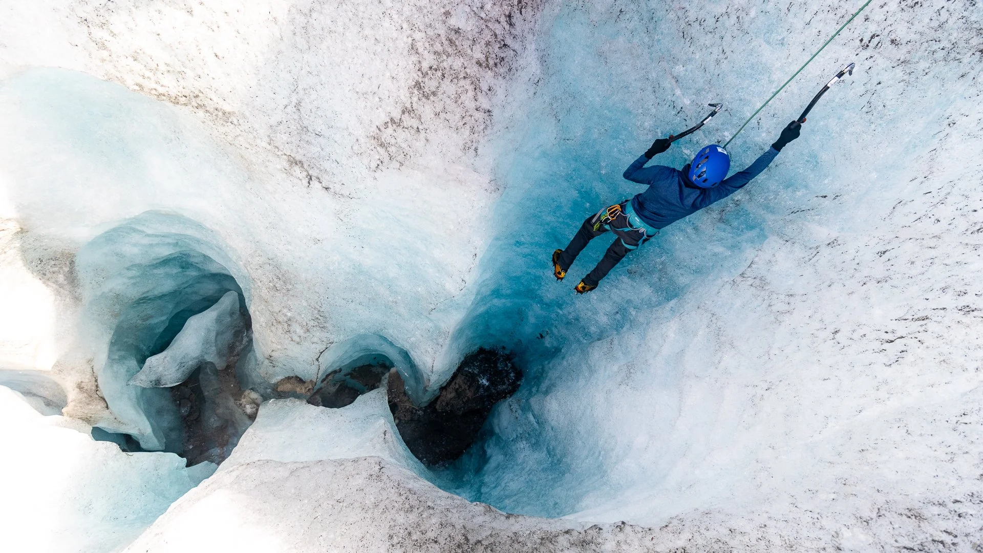 A person in blue winter gear, including a helmet and gloves, is rappelling into a crevasse in an icy glacier using an ice axe and safety harness.