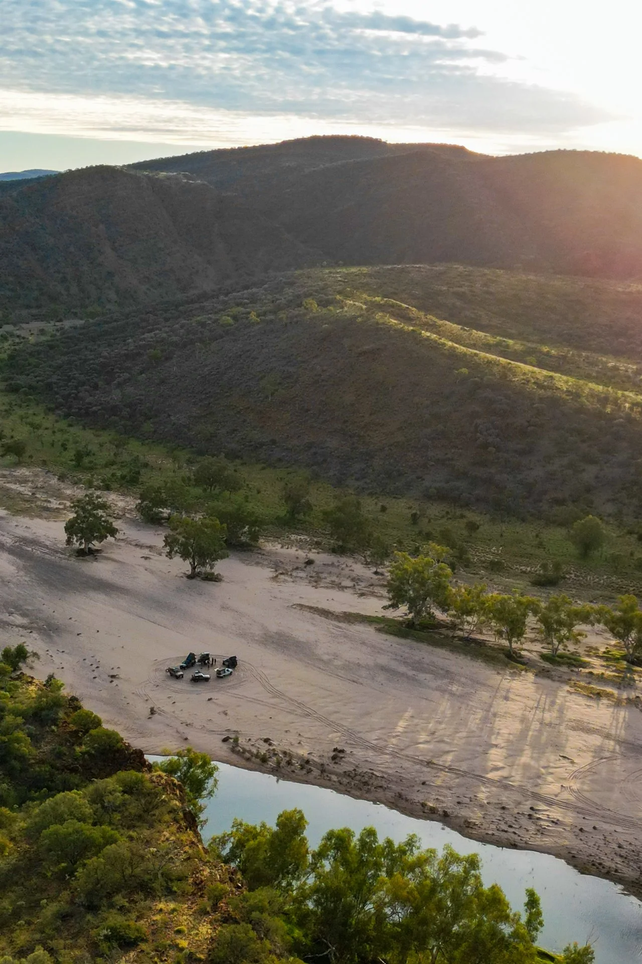 A view of mountains during sunset with a sandy area, trees, and a small body of water in the foreground. Several vehicles and people are gathered on the sandy area.