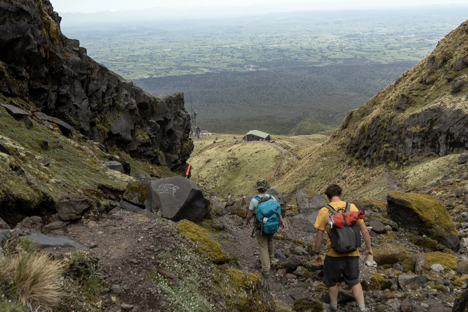 Hikers walking down a rocky mountain trail surrounded by grassy and moss-covered terrain with a mountain summit in the background. In the distance, a green building and communication tower are visible, overlooking a vast flat landscape.