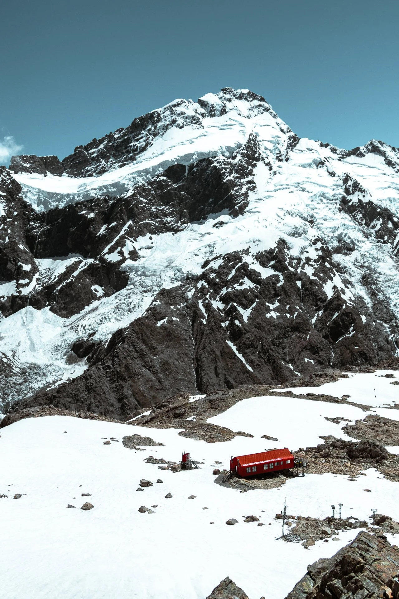 Snow-covered mountainous landscape with rocky peaks and a small red building at the base of the mountain.