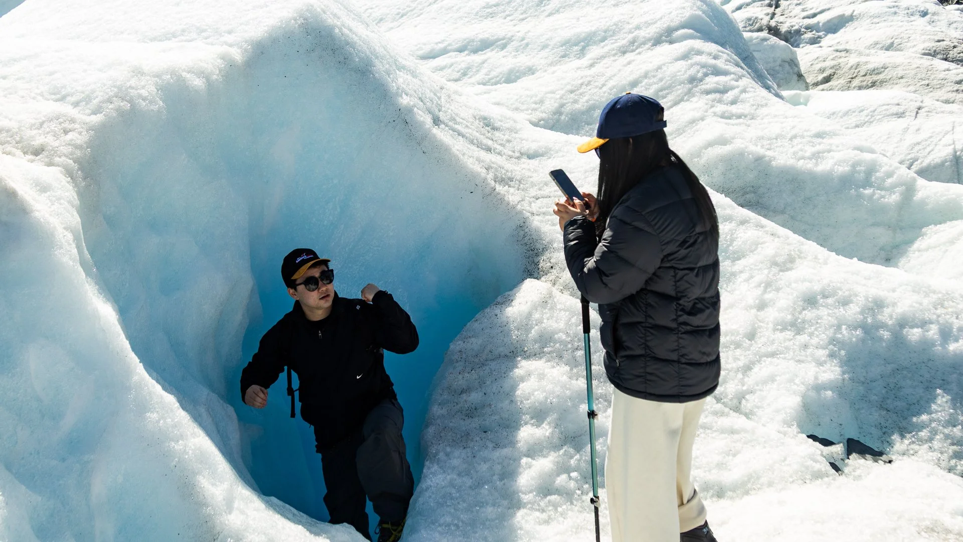 Two people explore an icy landscape with a crevasse, one person is standing and taking a photo while the other is stepping into the crevasse.