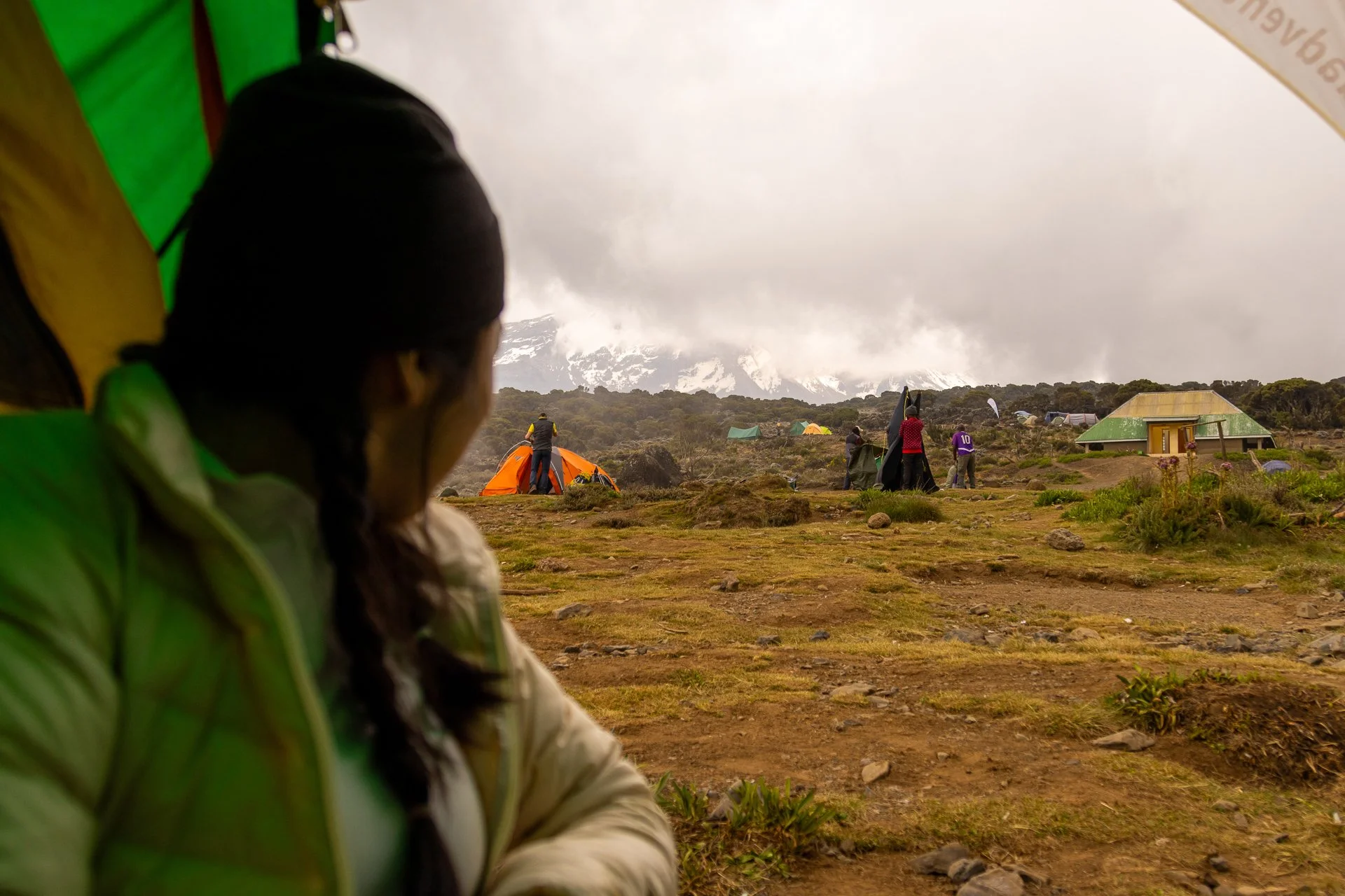 A group of people camping in a mountainous area with tents, some standing, some sitting, surrounded by clouds and snow-capped peaks.