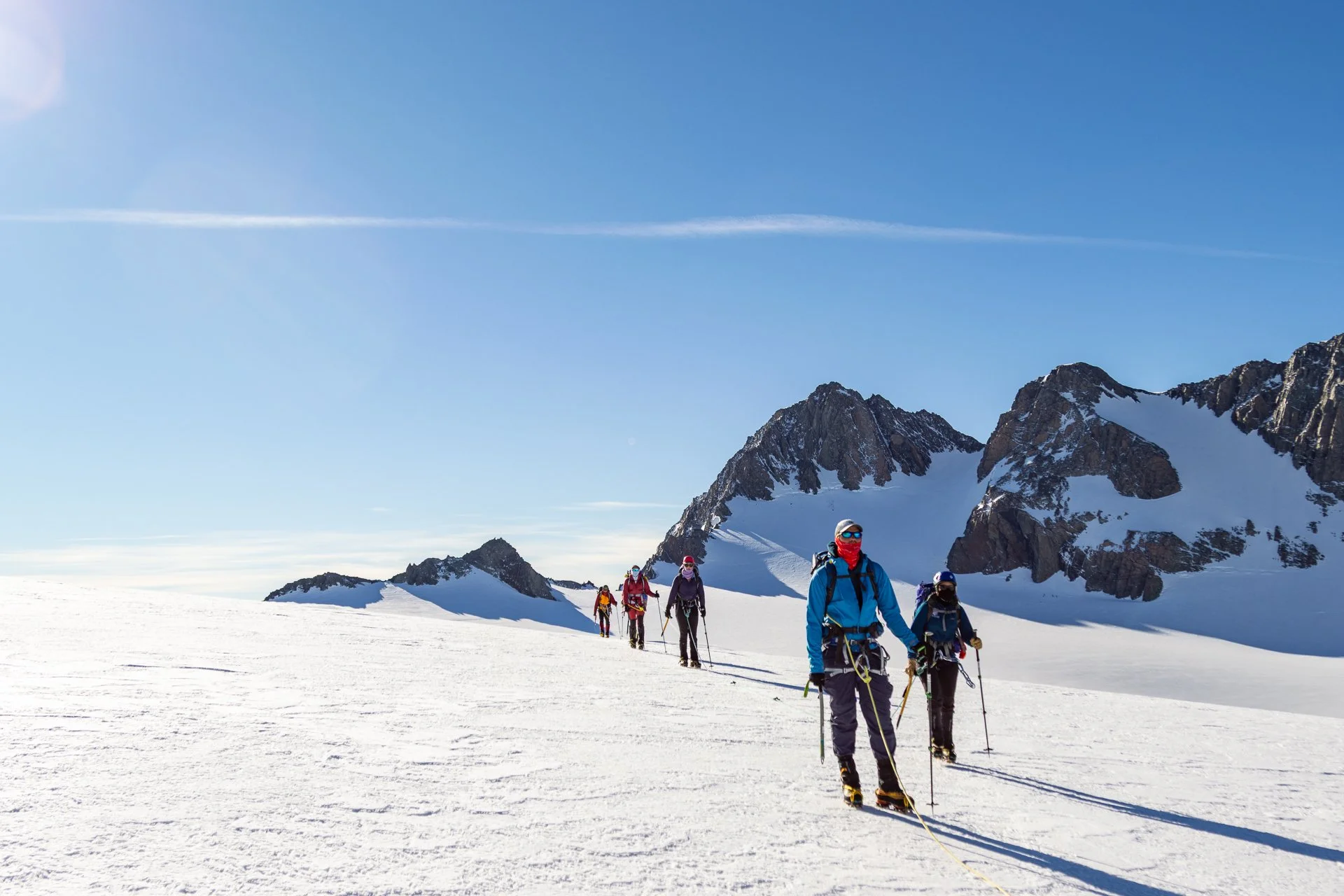 Group of skiers trekking across snow-covered mountains under a blue sky with rocky mountain peaks in the background.