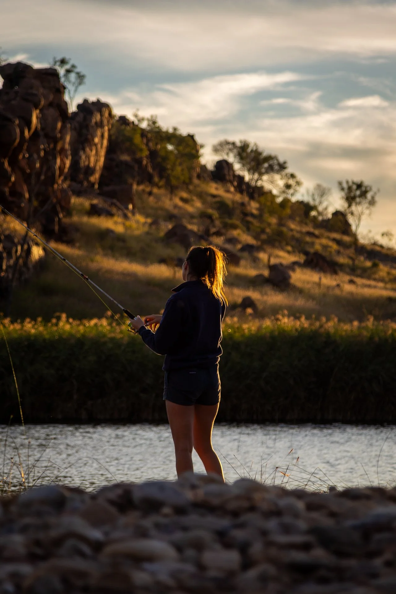 A person fishing by a river during sunset with rocky landscape and trees in the background.