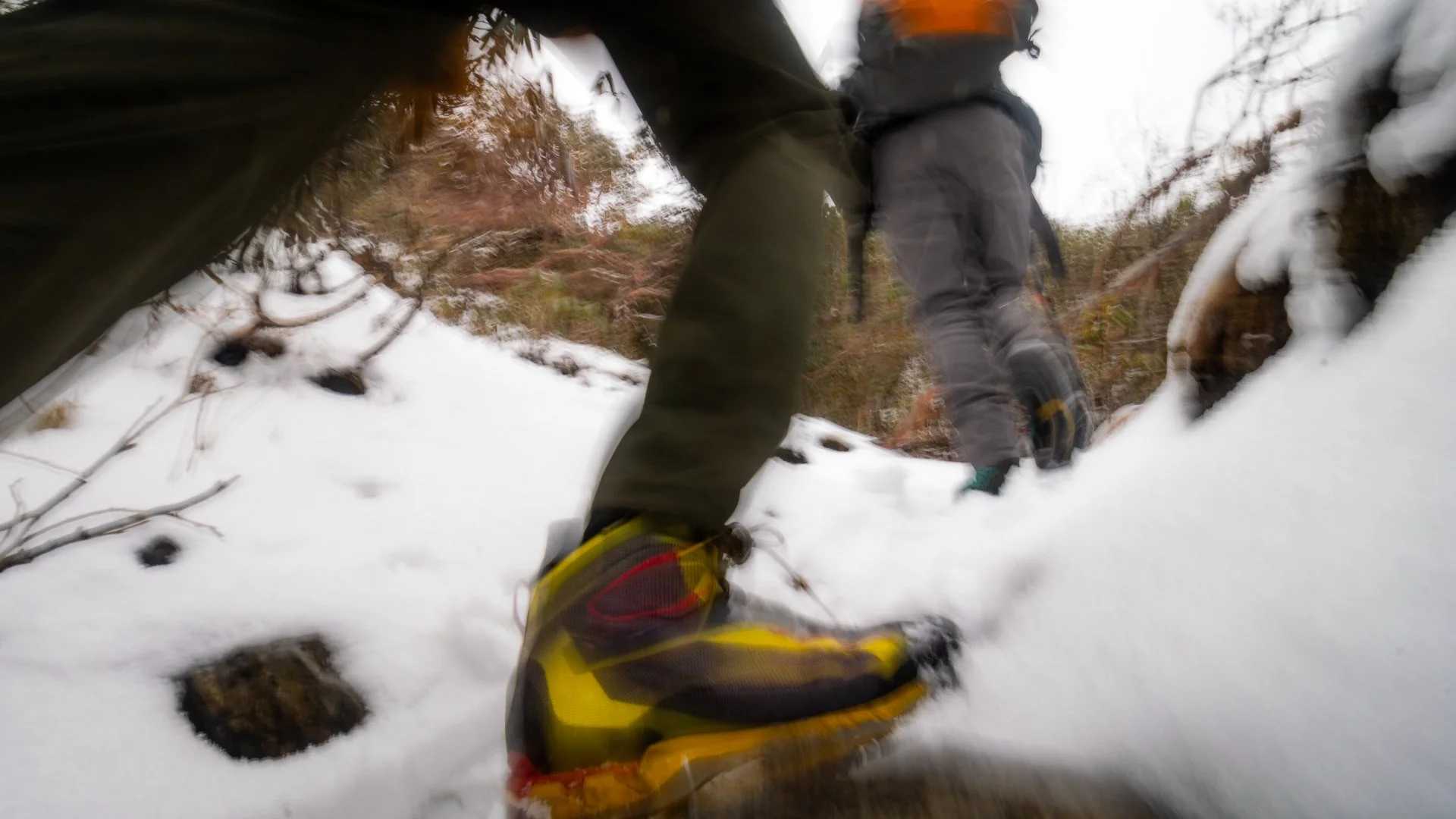 Person in black jacket and yellow boots falling or sliding in the snow in a wooded area, with another person in the background also in the snow.