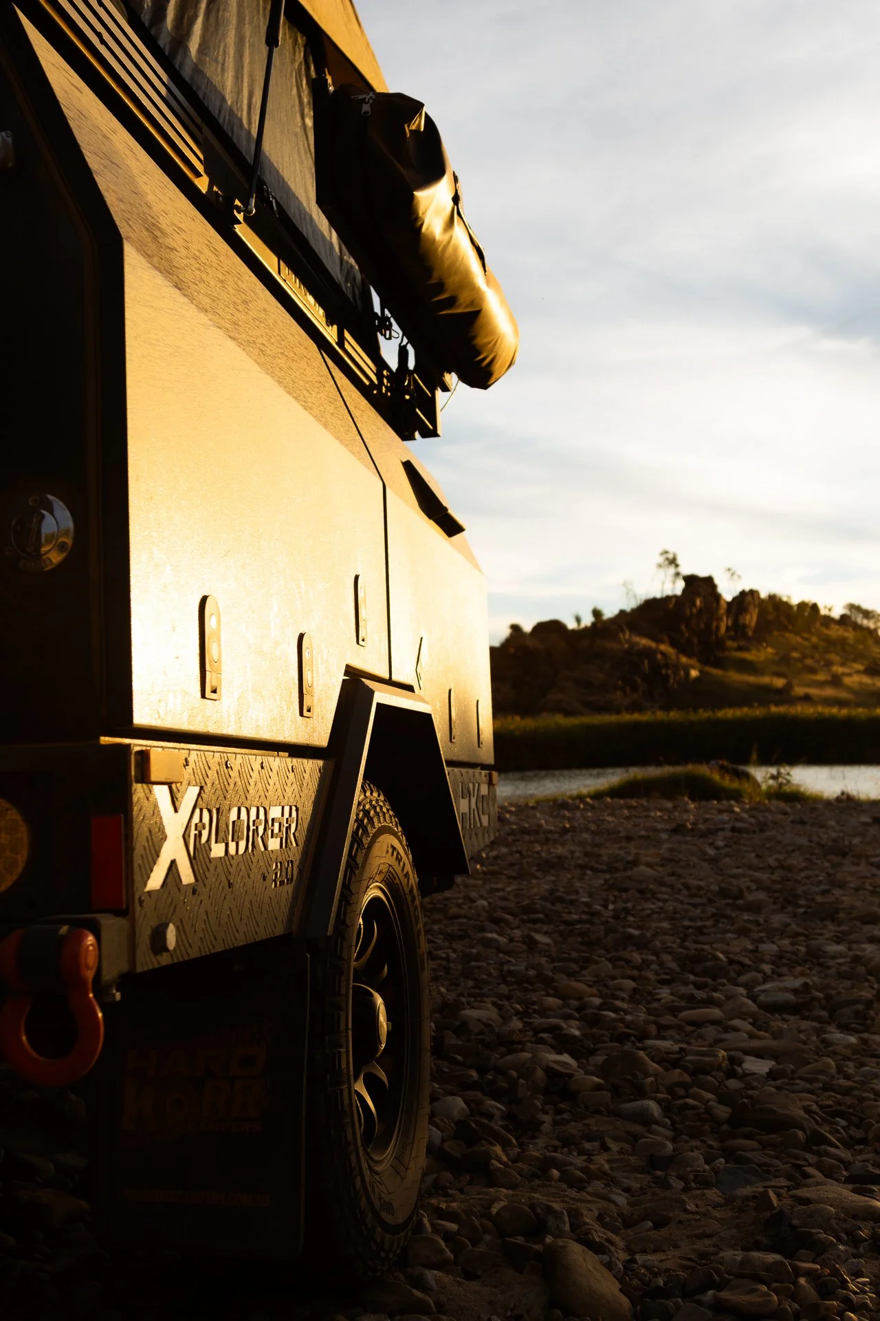 Close-up of a yellow off-road vehicle with equipment mounted on the roof, parked on a rocky riverbank during sunset, with hills and a river in the background.