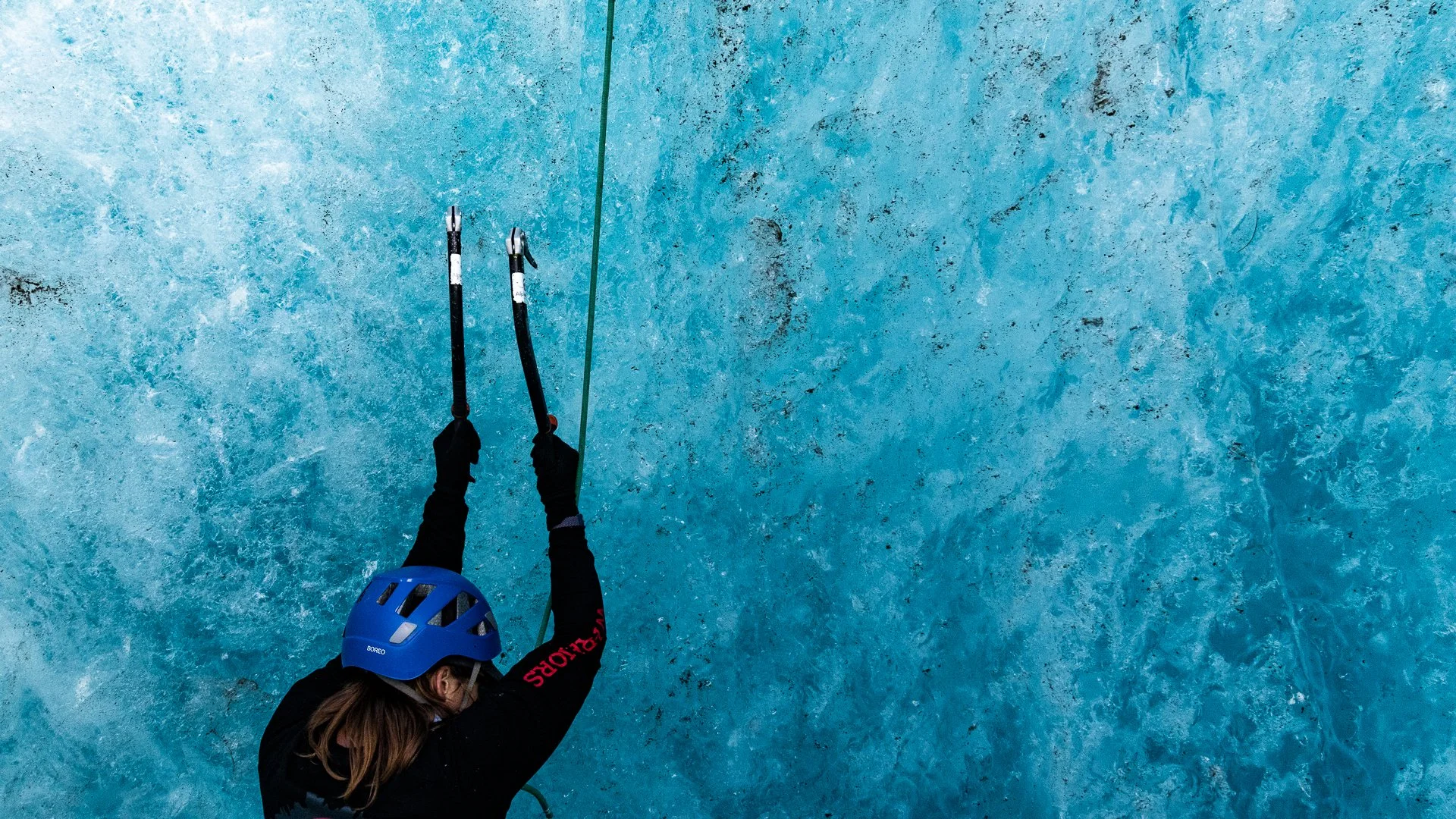 Person wearing a blue helmet and black gloves ice climbing on a blue ice wall using two ice axes.