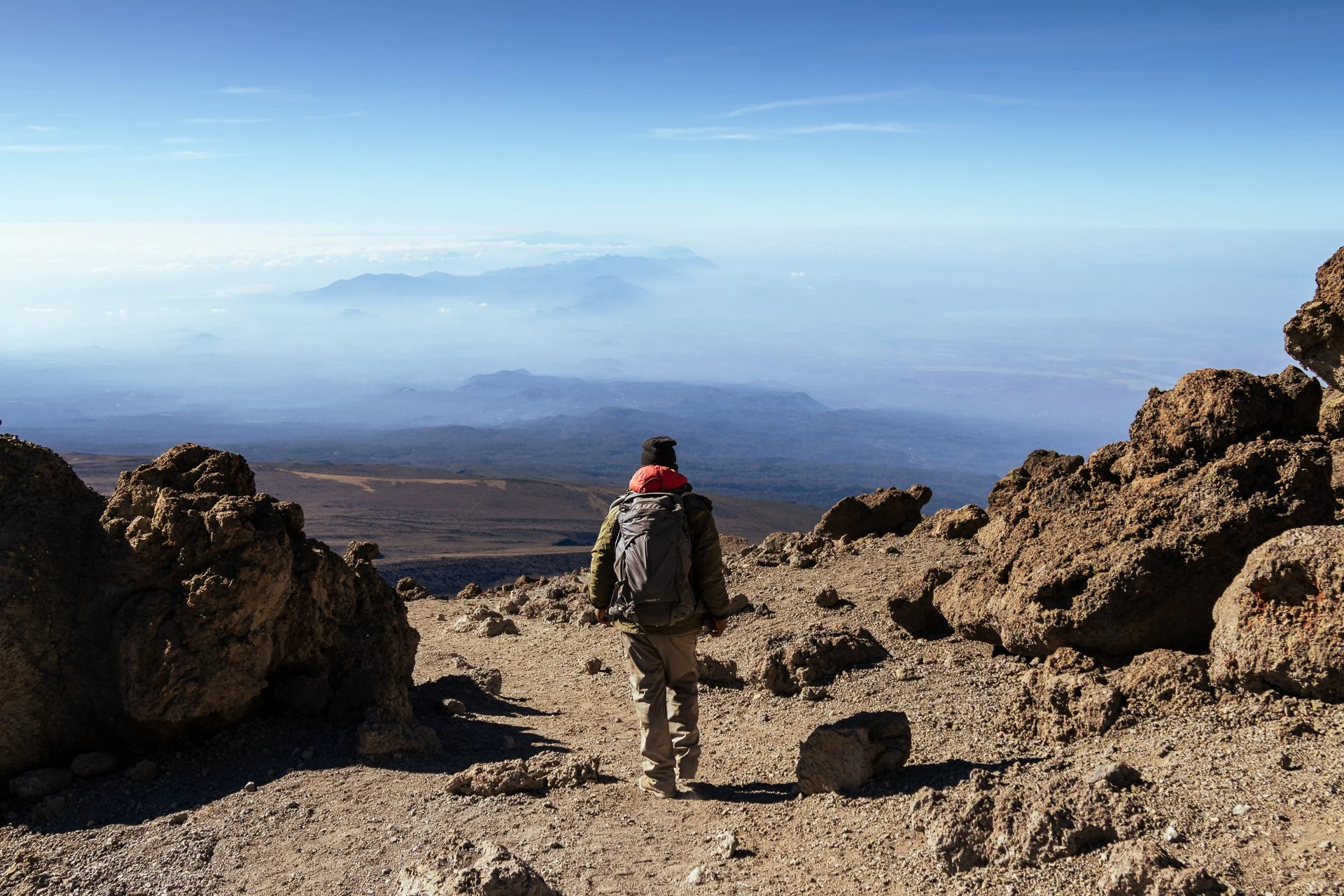 A person with a backpack hiking on rocky terrain in a mountainous area with a view of distant mountains and a cloudy sky.