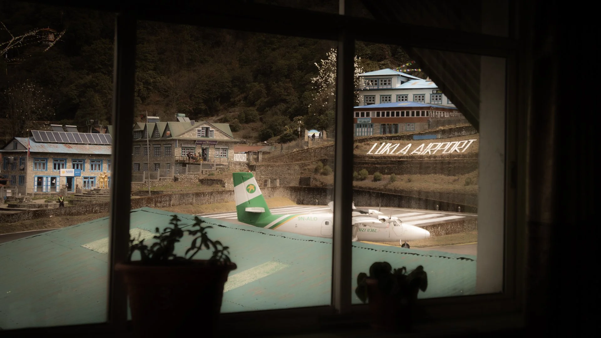 View of Lukla Airport with a white airplane on the runway, surrounded by buildings and a hillside with trees, seen through a window frame.