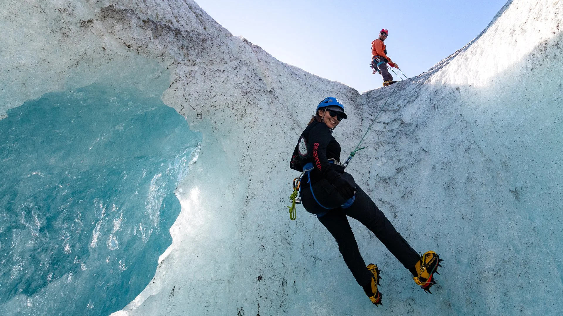 Two climbers descending a glacier with a crevasse, wearing helmets and harnesses, surrounded by ice and snow under a clear blue sky.