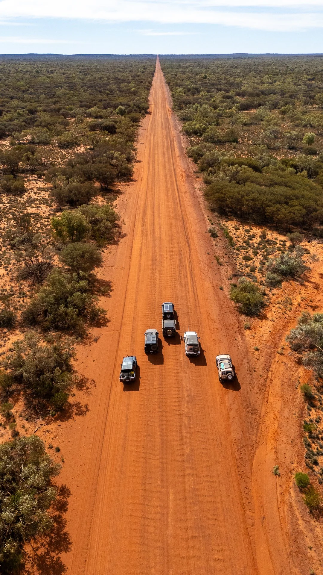 Six off-road vehicles driving on a long, straight, reddish dirt road through a sparse desert landscape with scattered bushes and trees under a partly cloudy sky.