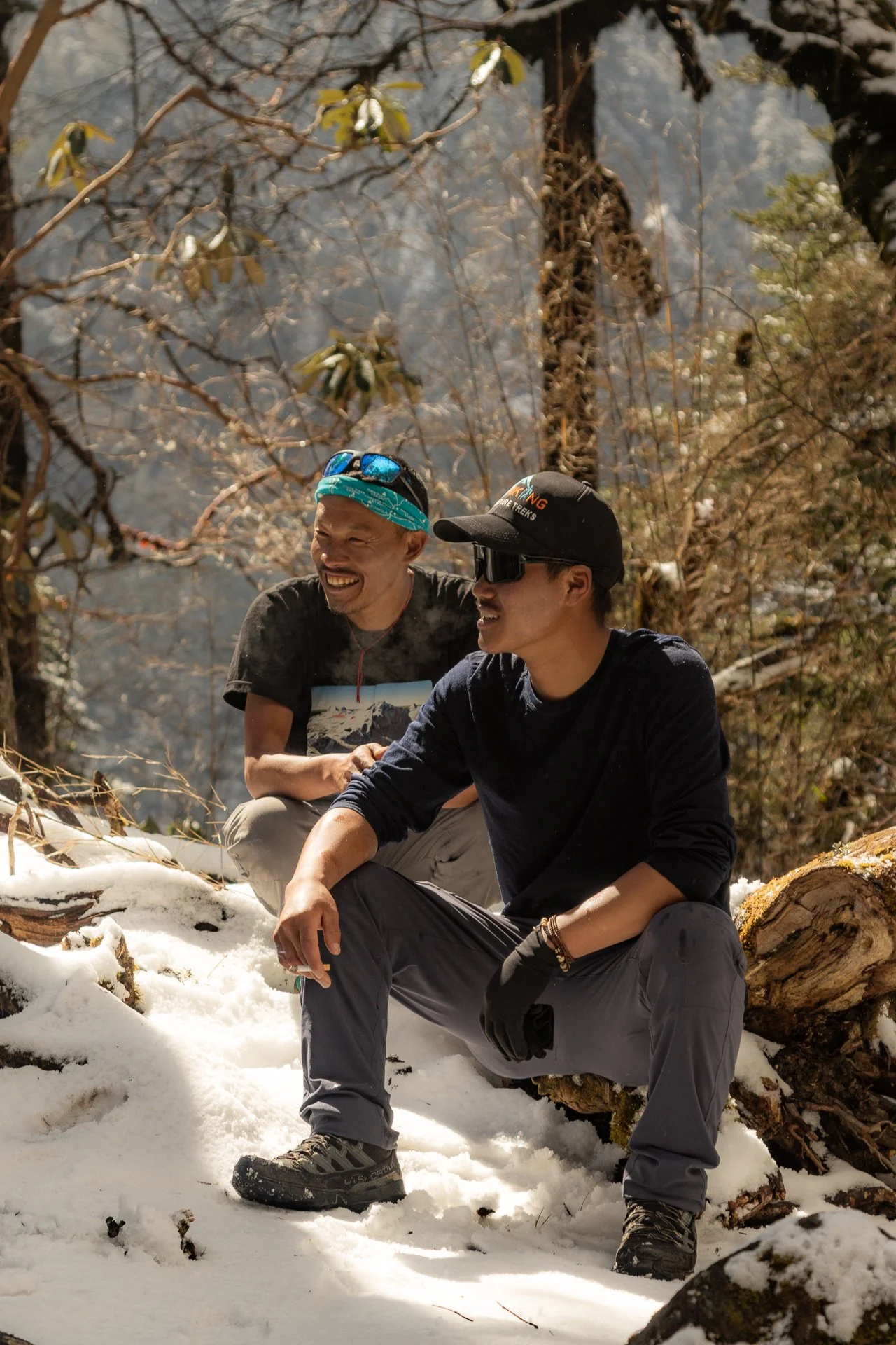 Two men sitting on snow-covered ground in a forest, smiling and talking, with trees and some snow on the branches surrounding them.