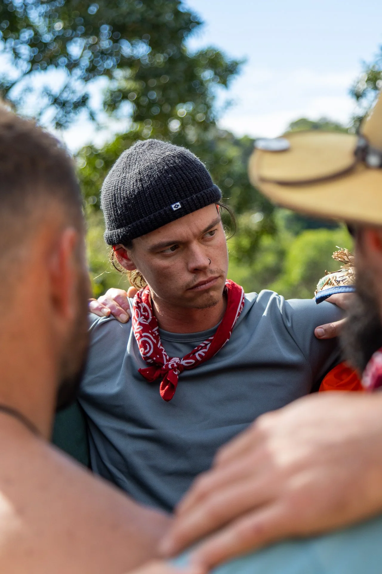 Group of people in a huddle outdoors, with a young man wearing a black beanie and red bandana around his neck in the center.